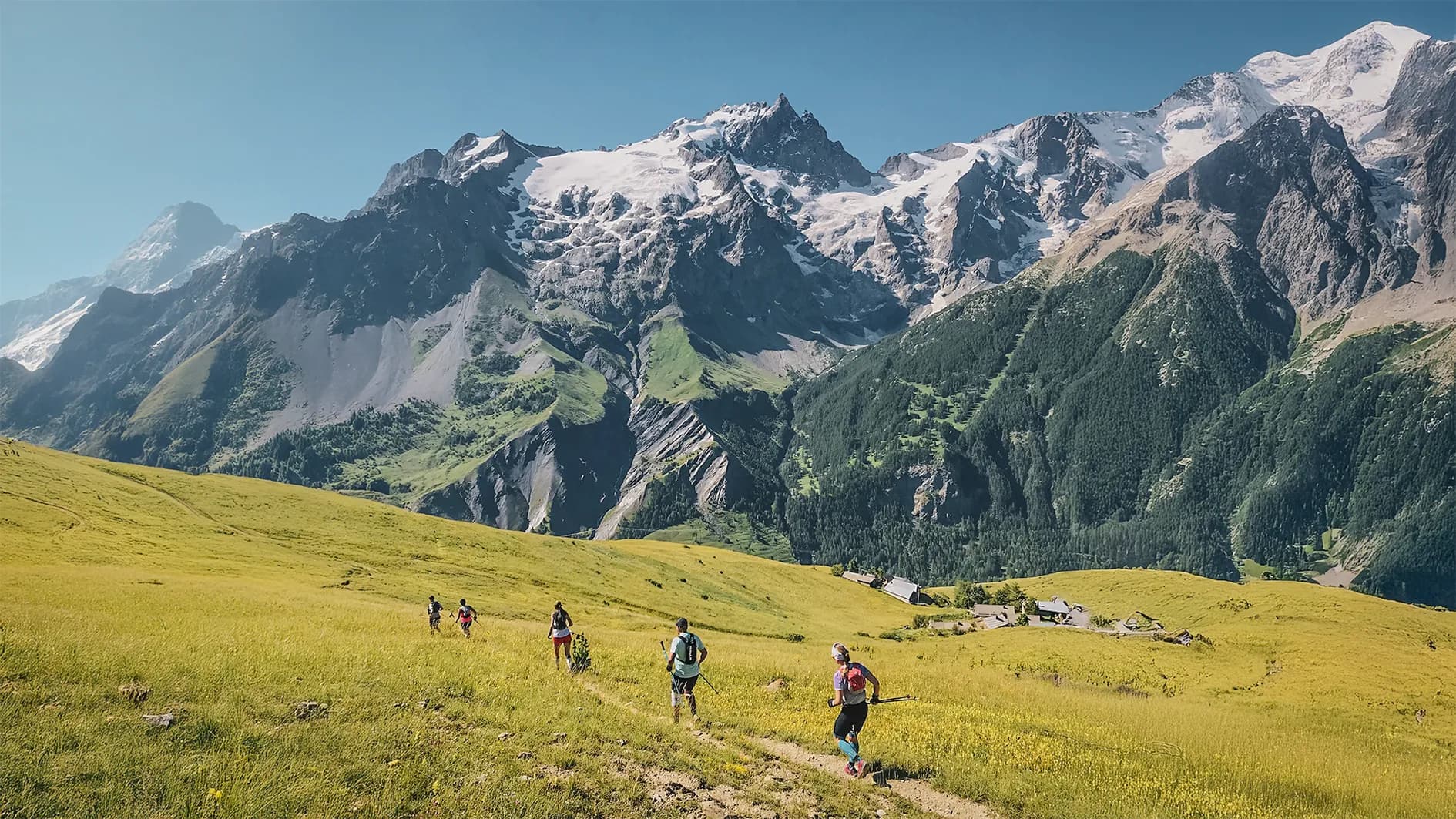 A group of hikers walk across a vast green meadow, surrounded by majestic snow-capped mountains. The hills are dotted with wildflowers and the sunlight illuminates the landscape.