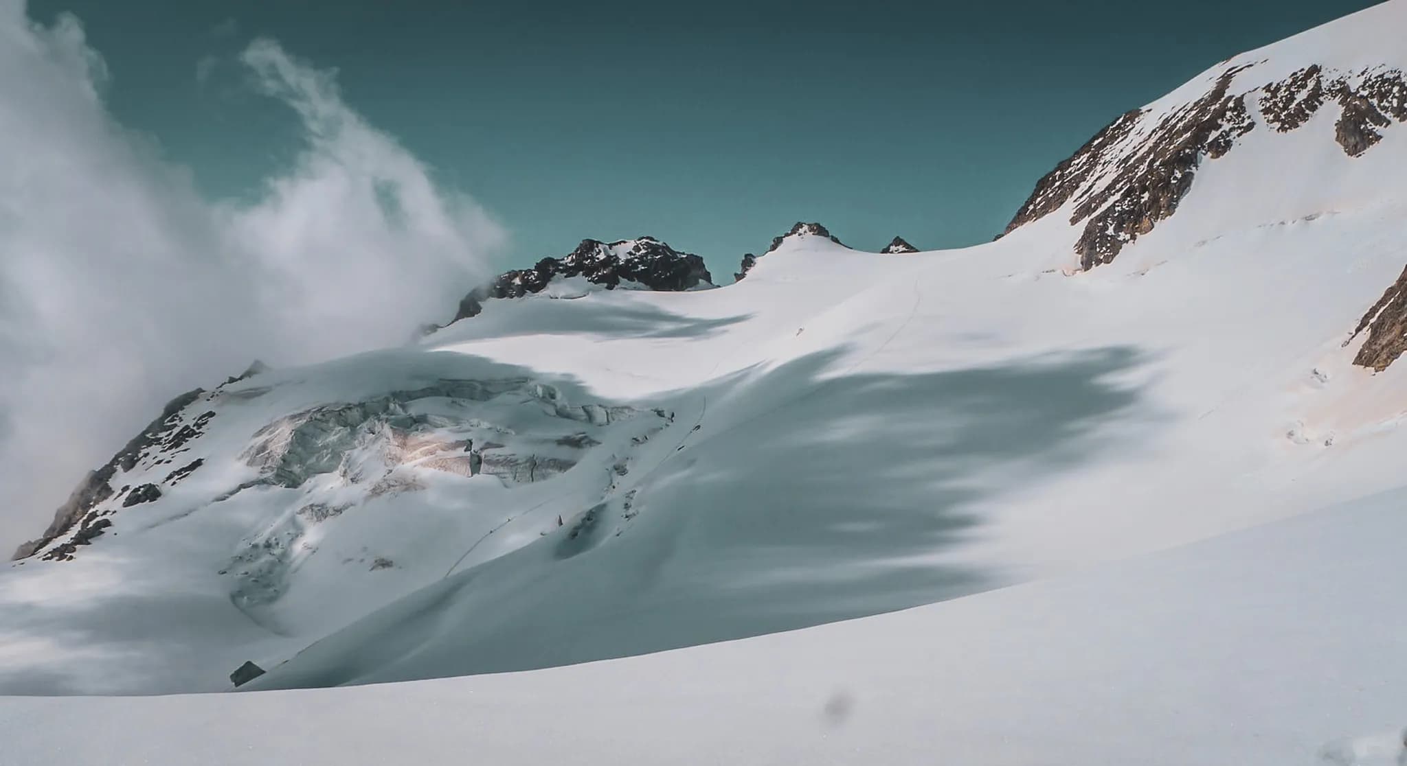 A majestic mountain landscape covered in snow. Steep peaks rise up in the background, dotted with dark rocks contrasting with the white immensity. Part of the glacier is visible, offering