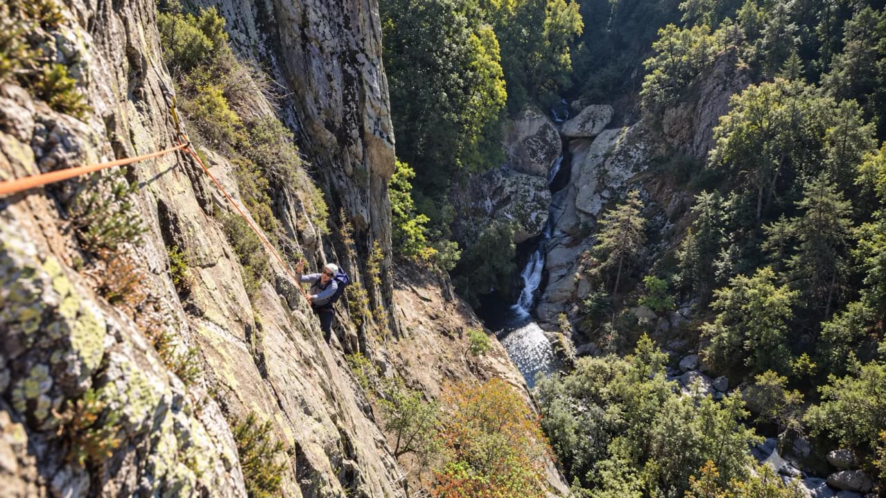 Een klimmer beklimt een rotswand die uitkijkt over een waterval, in het groene landschap van de Pyreneeën.