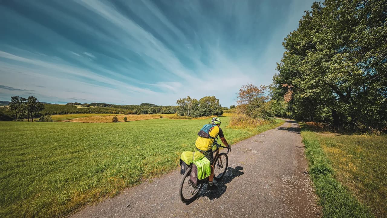 Cyclist on a country lane, surrounded by vast green meadows under a blue sky, inviting adventure.