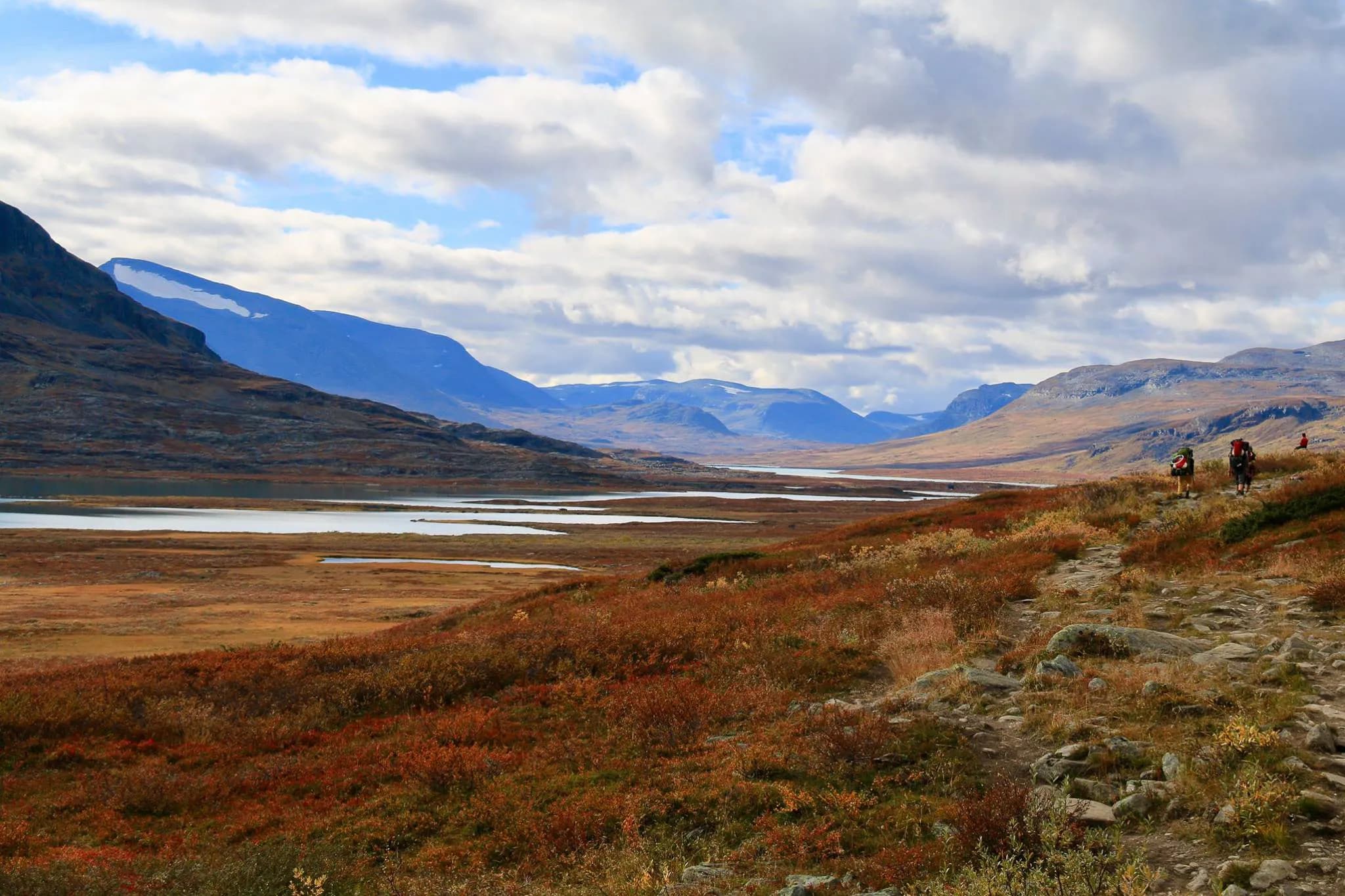 A mountainous landscape with majestic peaks under a cloudy sky. A vast lake is reflected in the valleys, surrounded by autumn-coloured lands, where reds and golds mingle.