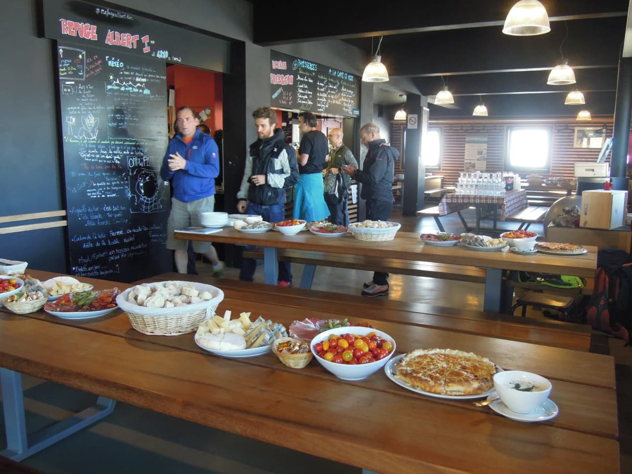 Gezellige ruimte met houten tafels, een verscheidenheid aan lokale specialiteiten en wandelaars op de achtergrond.