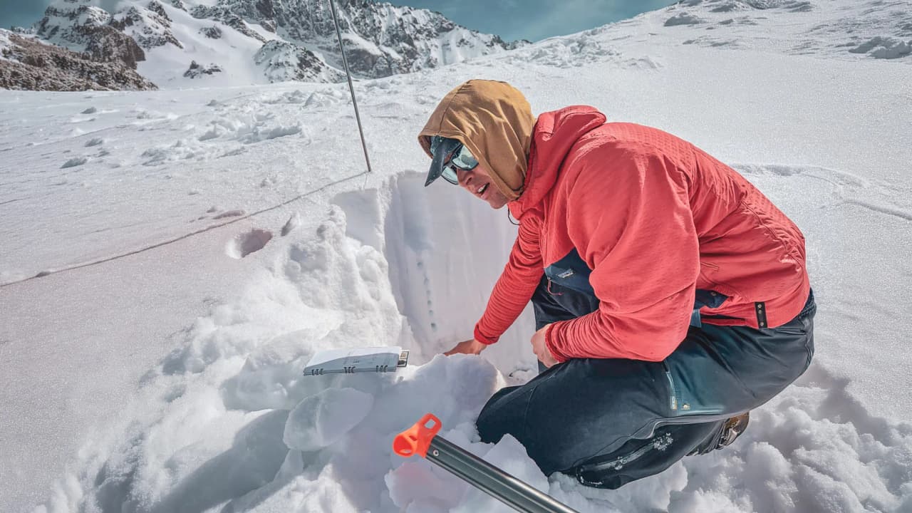 A skier examines the snow, surrounded by majestic Alpine peaks under a clear sky.