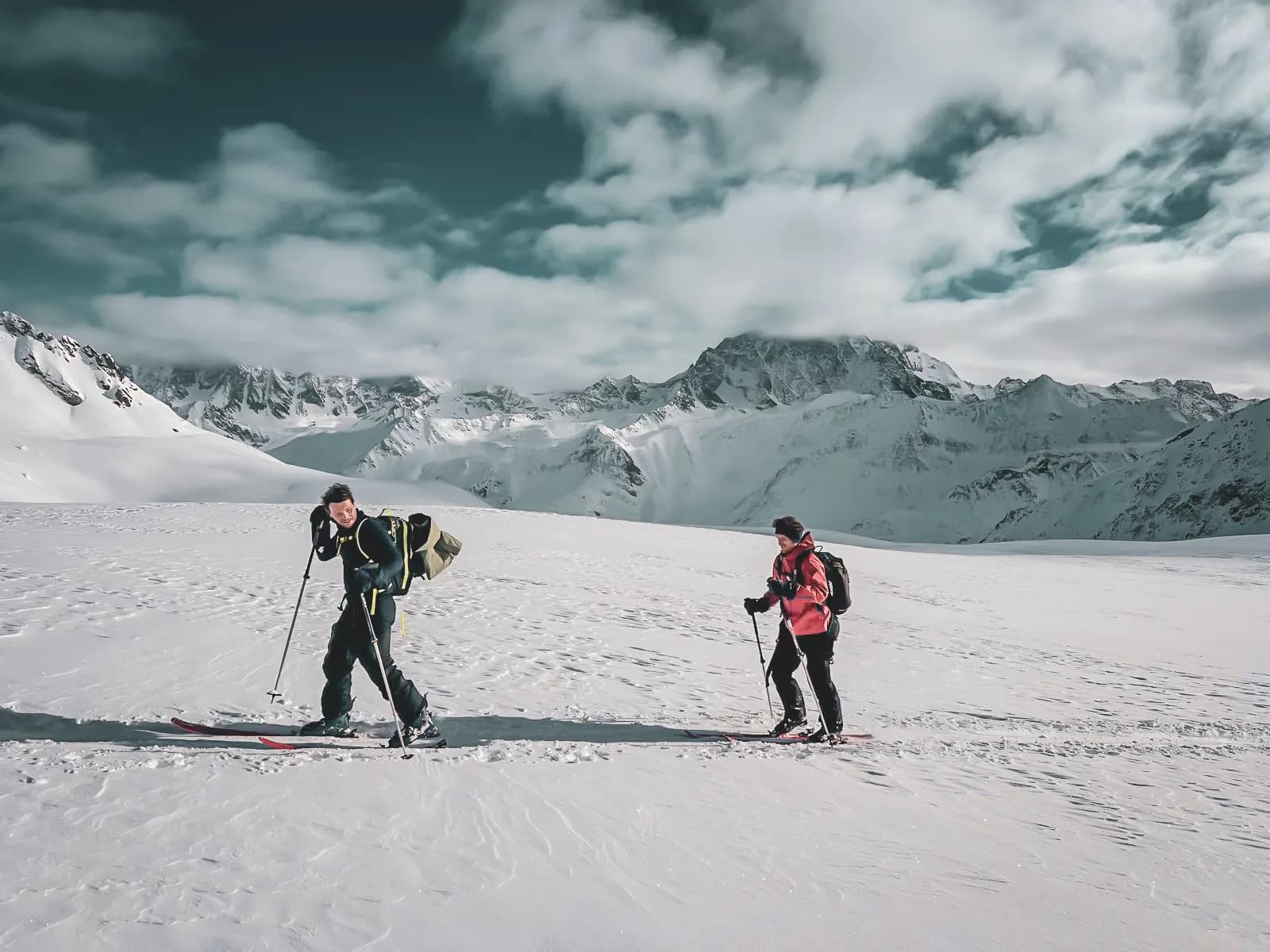 A snowy mountain landscape with two skiers making their way through the snow. One of the skiers is wearing a rucksack and ski equipment, while the other is wearing a red jacket,