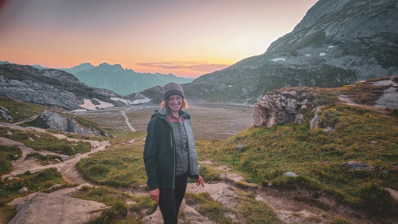 A smiling hiker in a majestic Alpine landscape, with mountains and colourful skies at sunset.