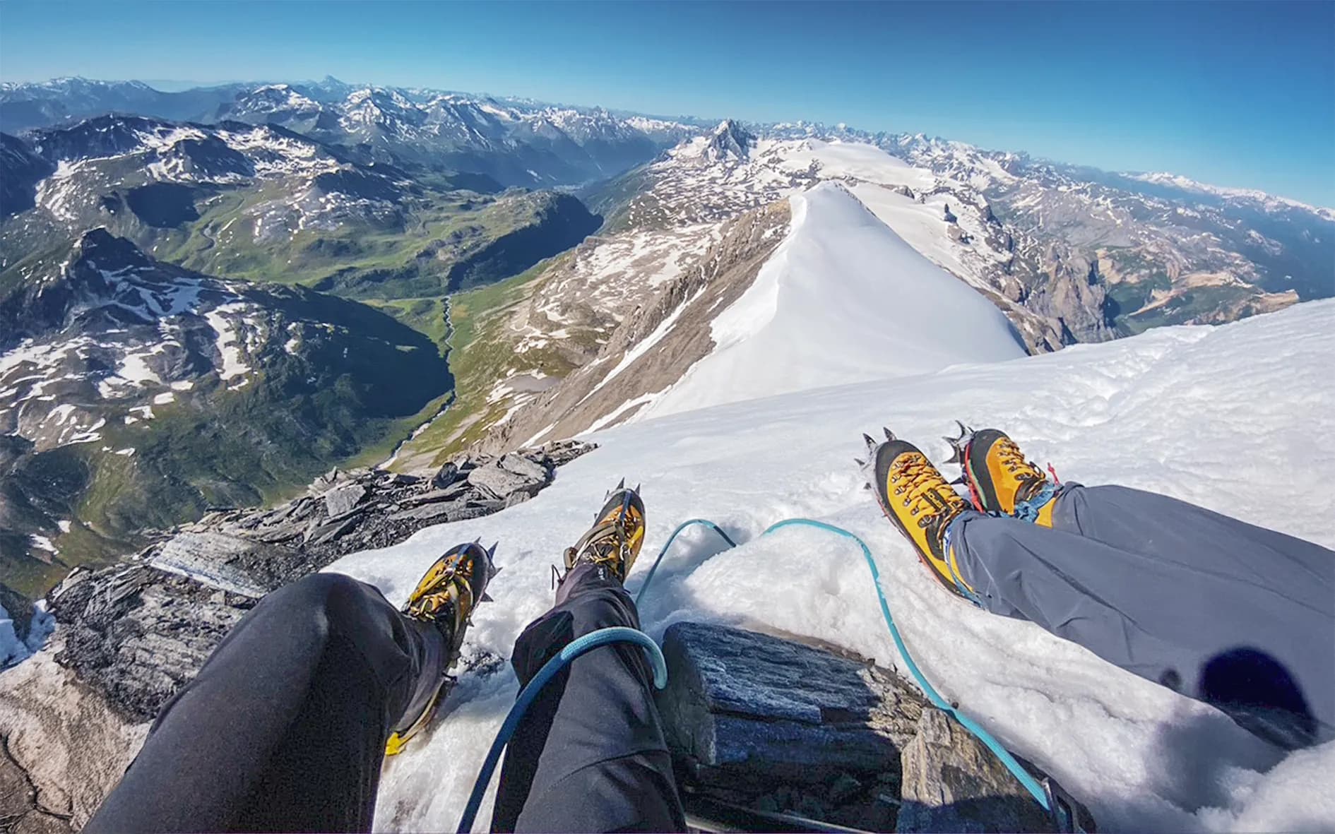 A group of three climbers are out on a glacier under a clear sky. They are wearing climbing gear and crampons, with ropes binding them together. In the background, majestic mountains and