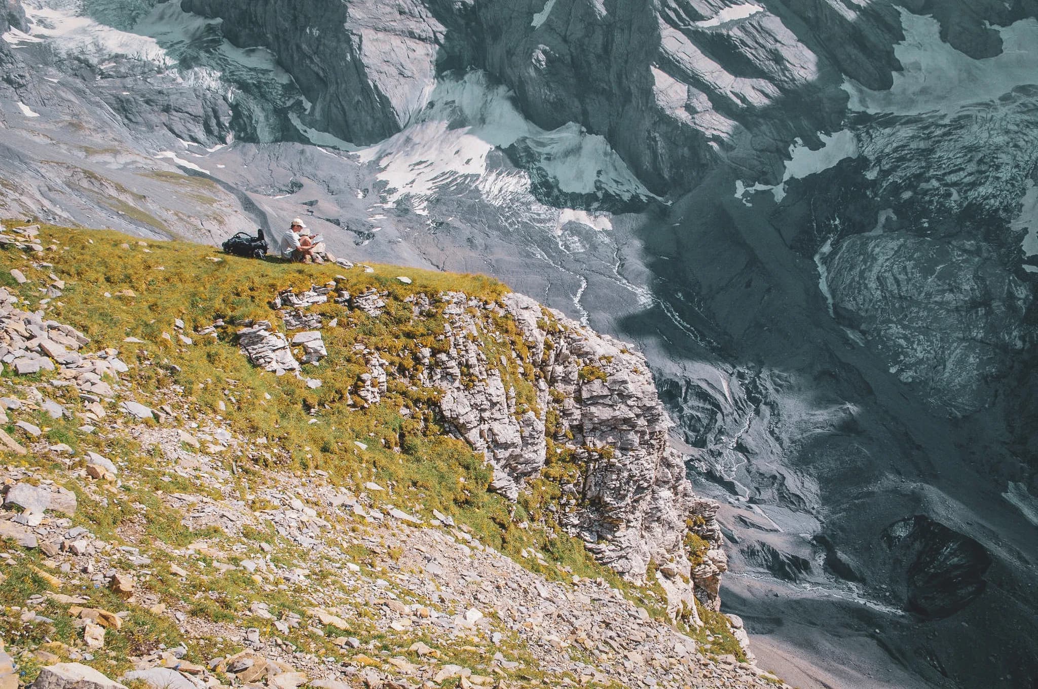 A spectacular mountain landscape with a bird's-eye view of rocky cliffs and glaciers. In the foreground, grassy, stony terrain with a few wild flowers.
