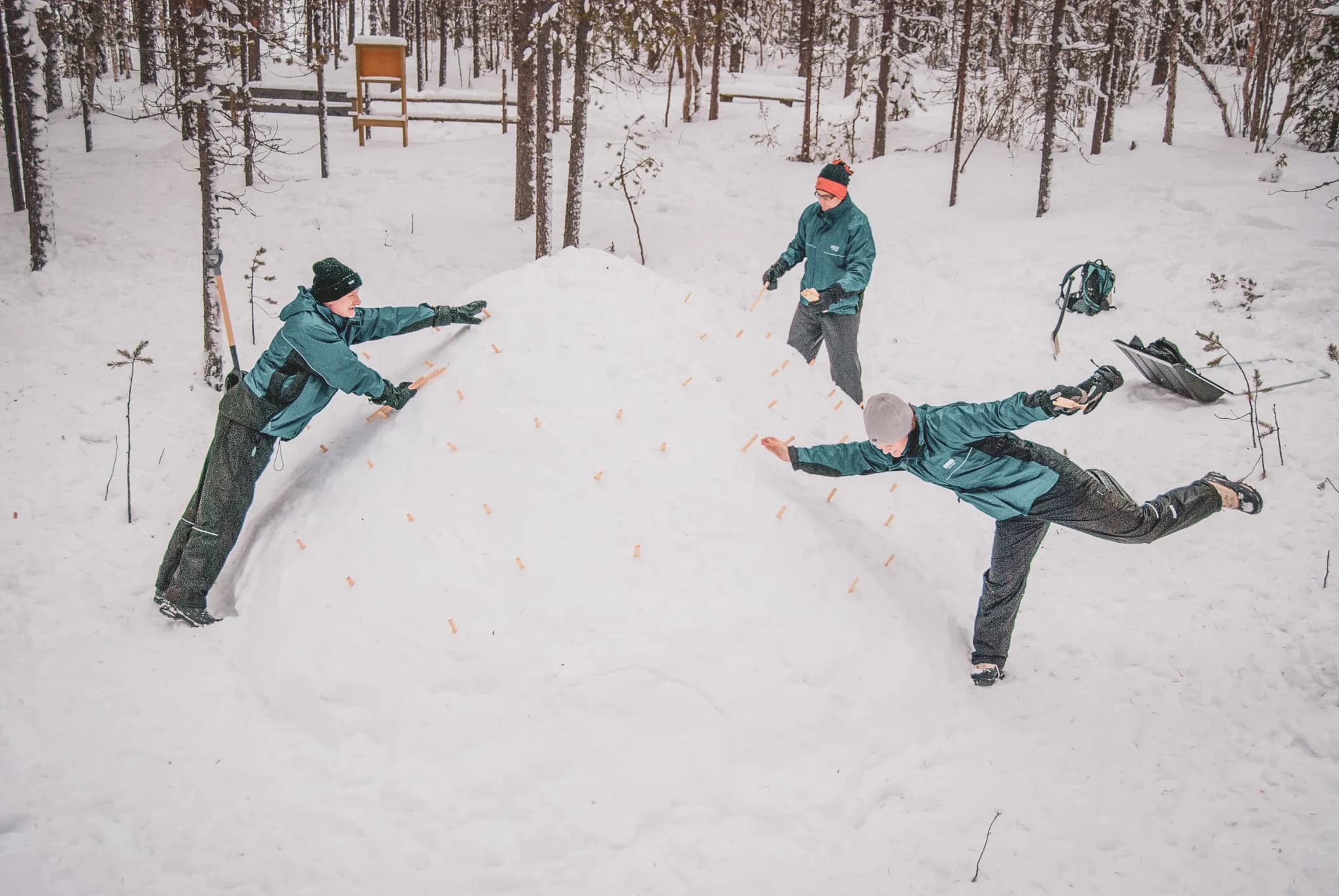 A group of people in winter clothes work together to build an igloo in the snow. The scene takes place in a forest, surrounded by trees, with a thin layer of snow covering the ground. Three people are busy