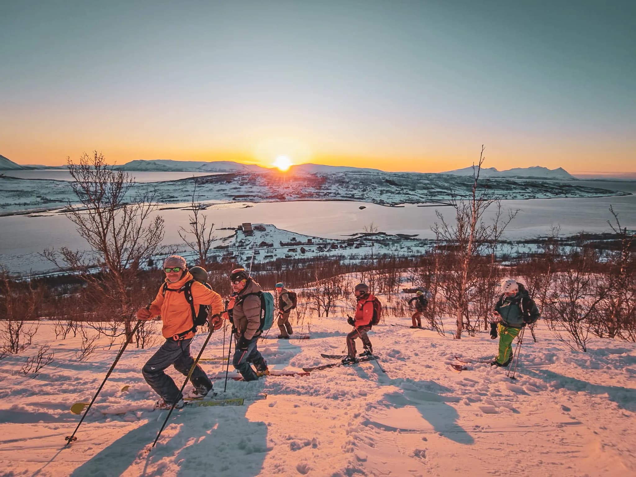 Un groupe de skieurs gravit une pente enneigée avec des arbres dispersés autour d'eux. Au fond, un magnifique paysage de montagnes se déploie sous un ciel clair où le soleil se lève, projetant une lumière dorée