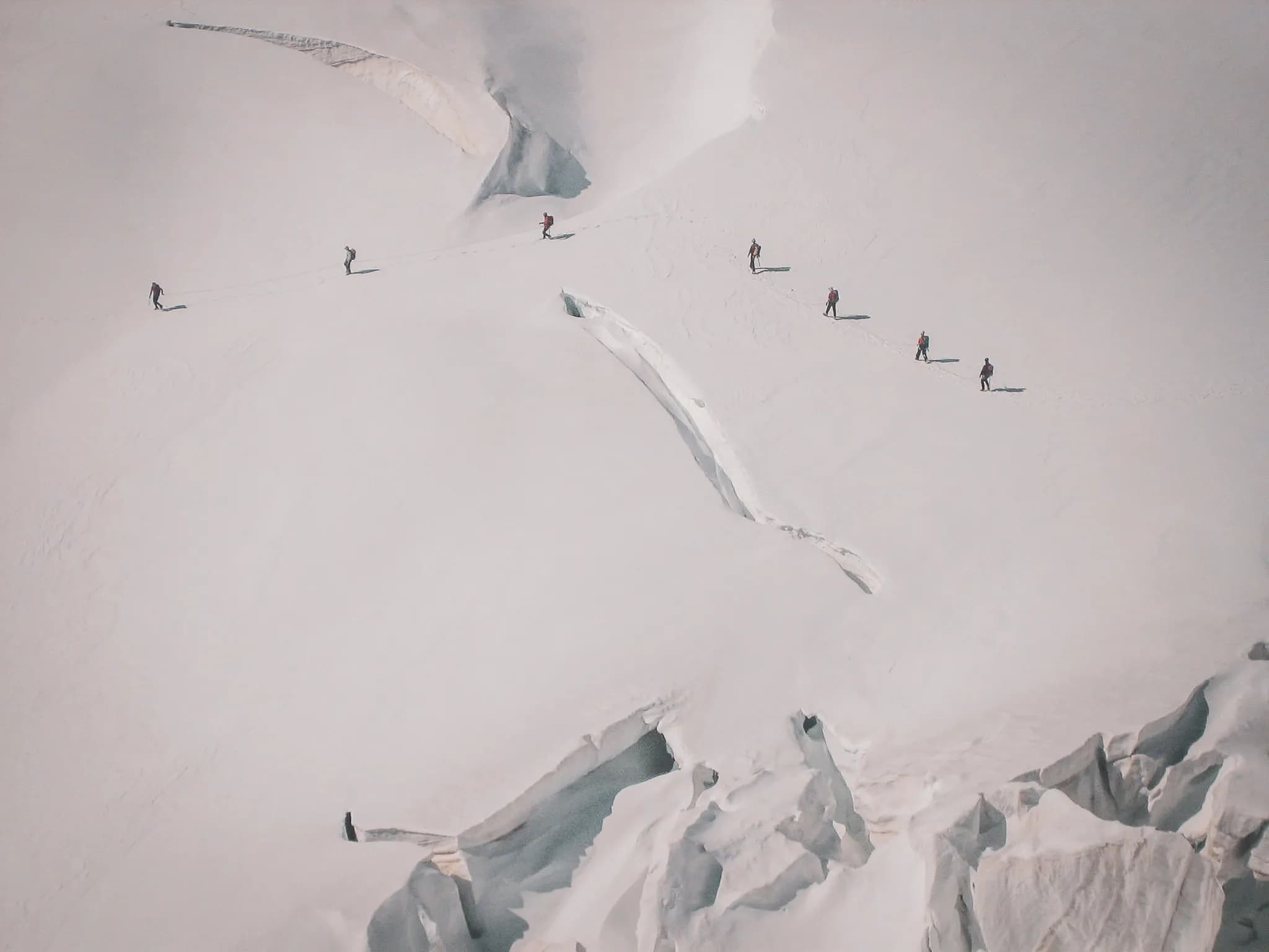 A glacial landscape stretches as far as the eye can see, with vast expanses of immaculate snow. Several climbers, dressed appropriately for the cold, are advancing in a line.
