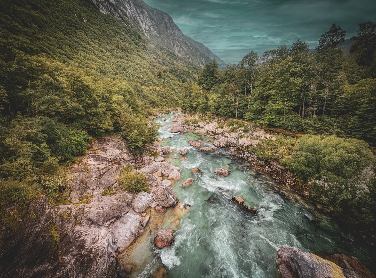 A turquoise stream winds through a rocky canyon, framed by sheer cliffs. In the basin, several people in orange canoes, dressed in life jackets, are paddling along.
