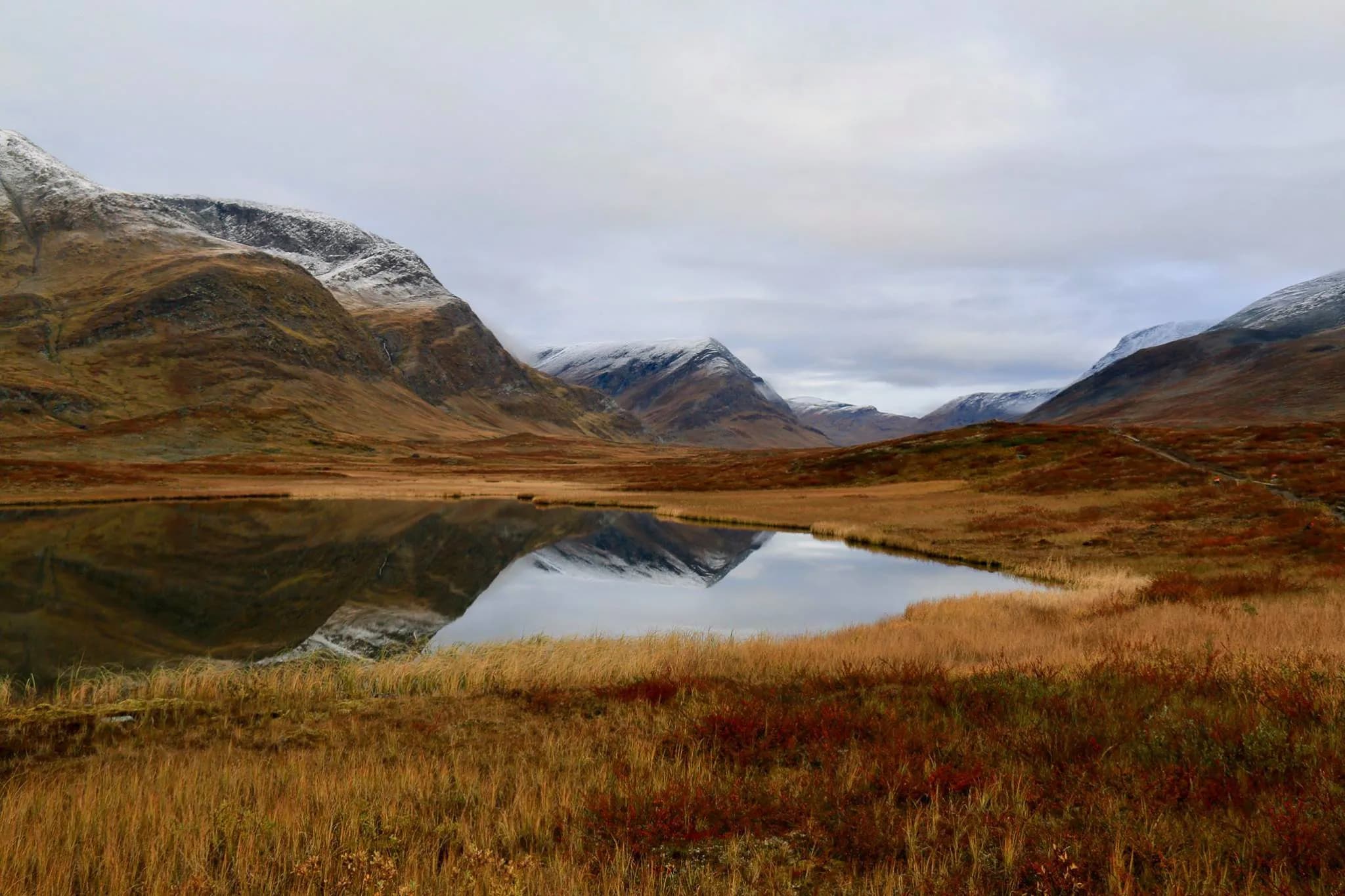 A mountainous landscape at sunset, with snow-capped peaks and a vast plain covered in golden grass. A calm lake reflects the surrounding mountains, creating a harmony of natural elements.