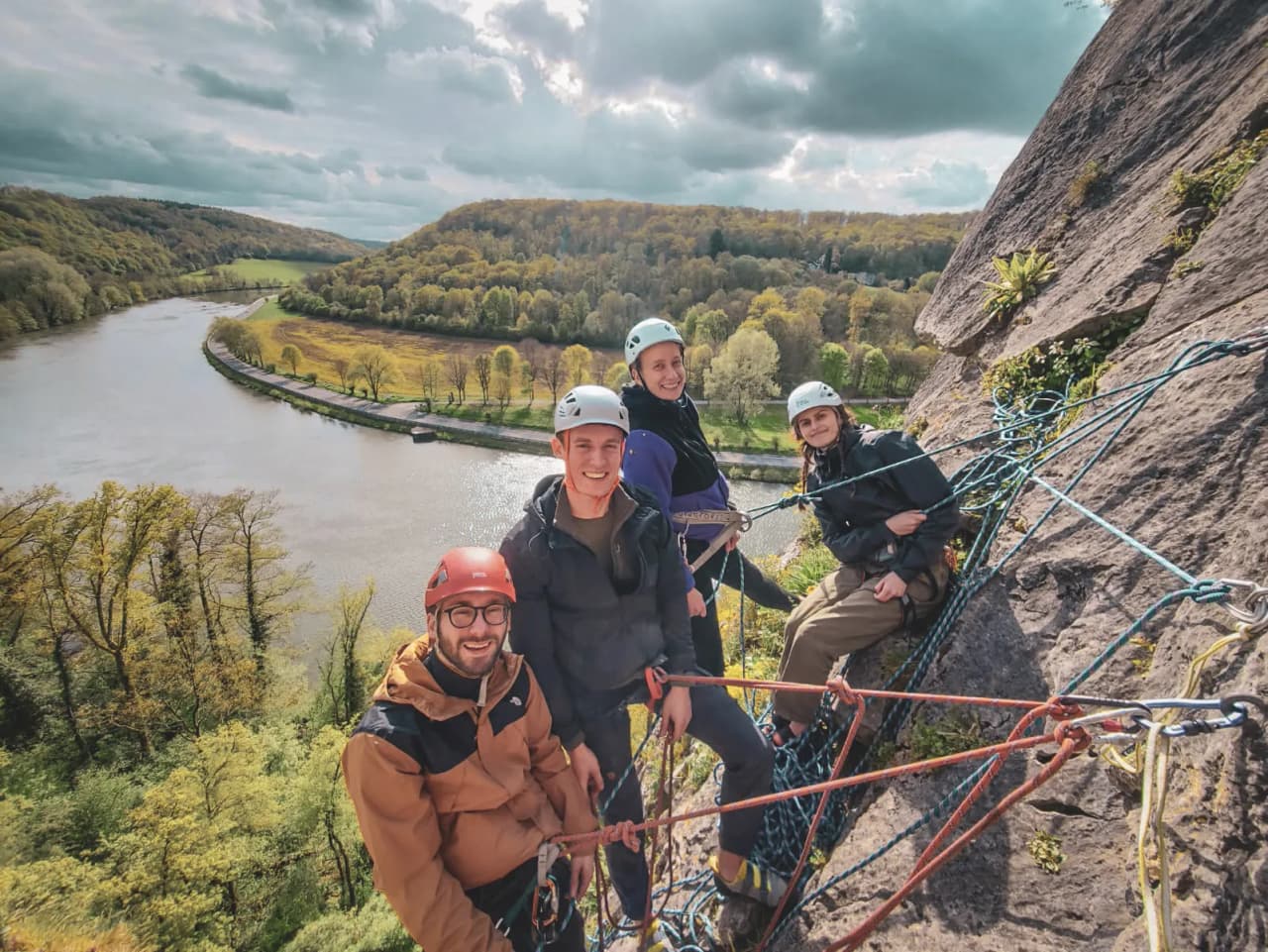 Quatre grimpeurs souriants posent sur une falaise surplombant une belle vallée verdoyante.