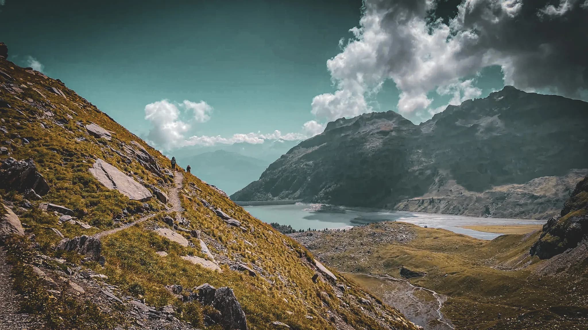 A majestic mountain landscape with a steep hill lined with yellow grass and rocks. A winding path crosses the slope, leading to a spectacular view of a lake below, surrounded by a forest.