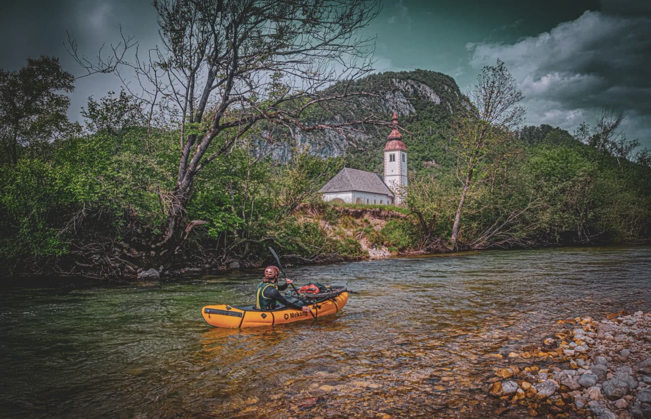 An orange kayak is paddling on a calm river, with one person on board wearing a helmet and wetsuit. In the background is a white church with a red steeple, surrounded by greenery. Mountains