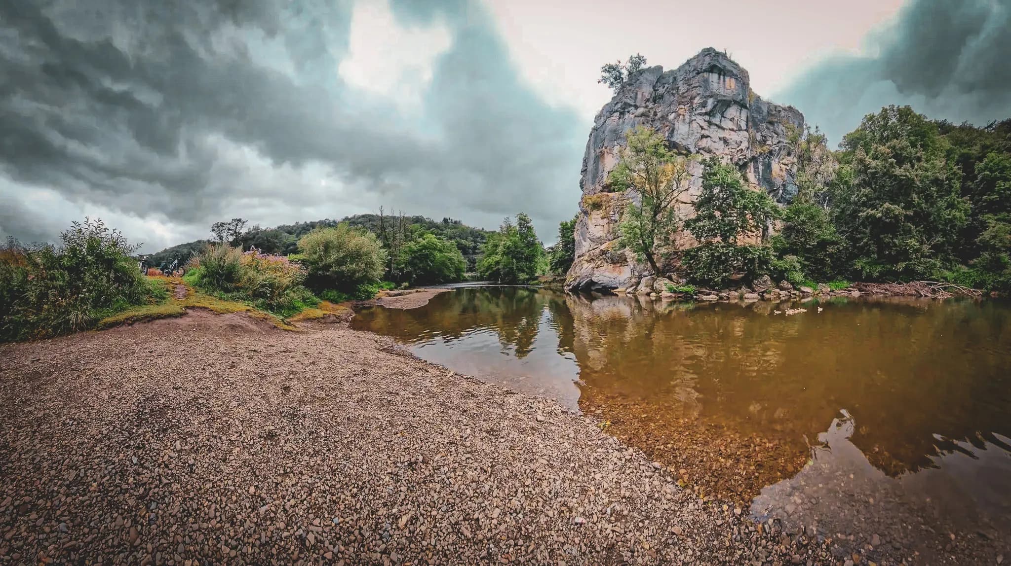 A natural landscape with a large craggy rock overlooking a calm stream. The sky is cloudy, with grey tones, and the surrounding vegetation is dense with trees and bushes. At the first