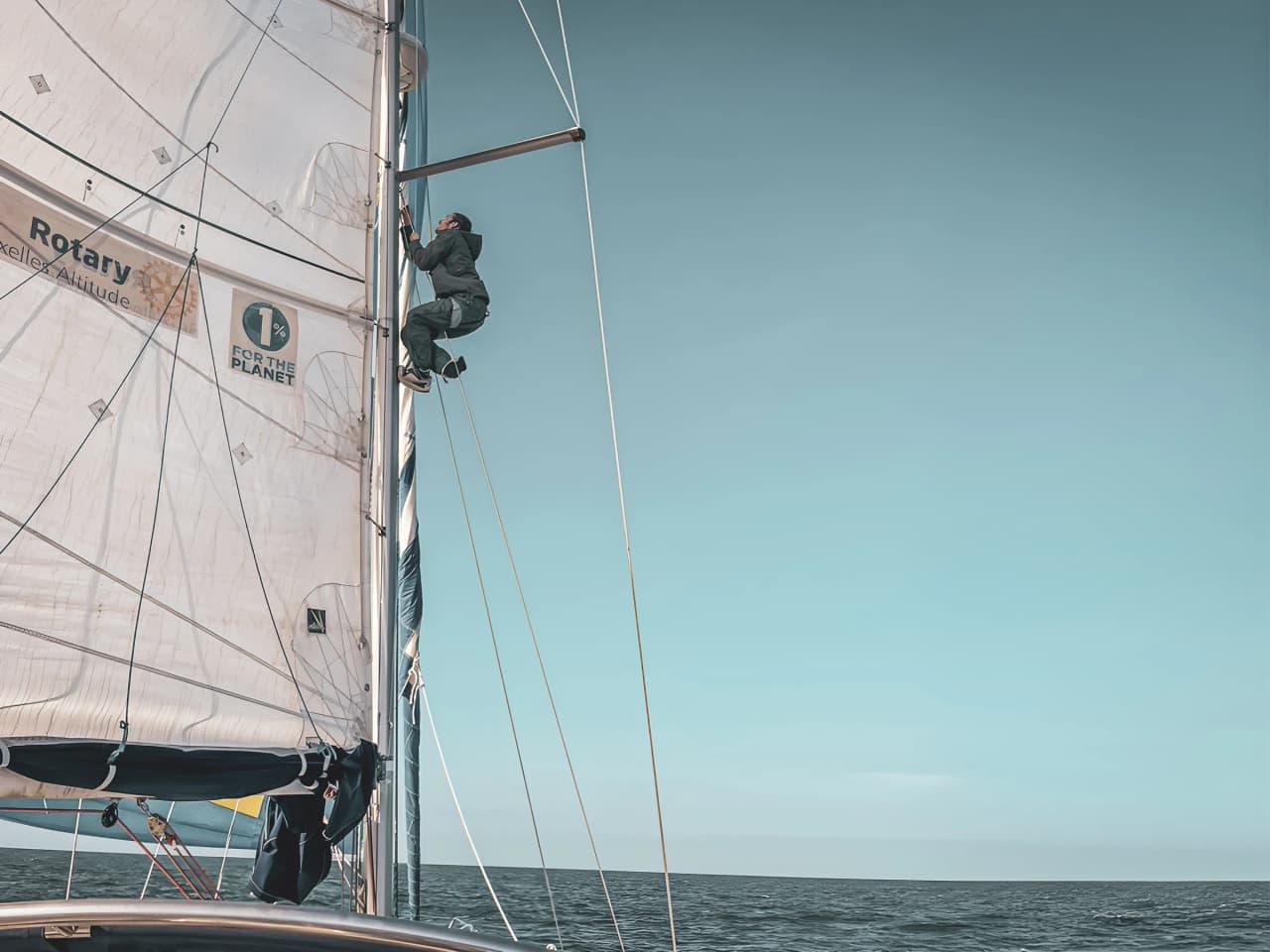 Un marin grimpe sur le mât d’un voilier, sous un ciel bleu, explorant les côtes du Pays de Galle.