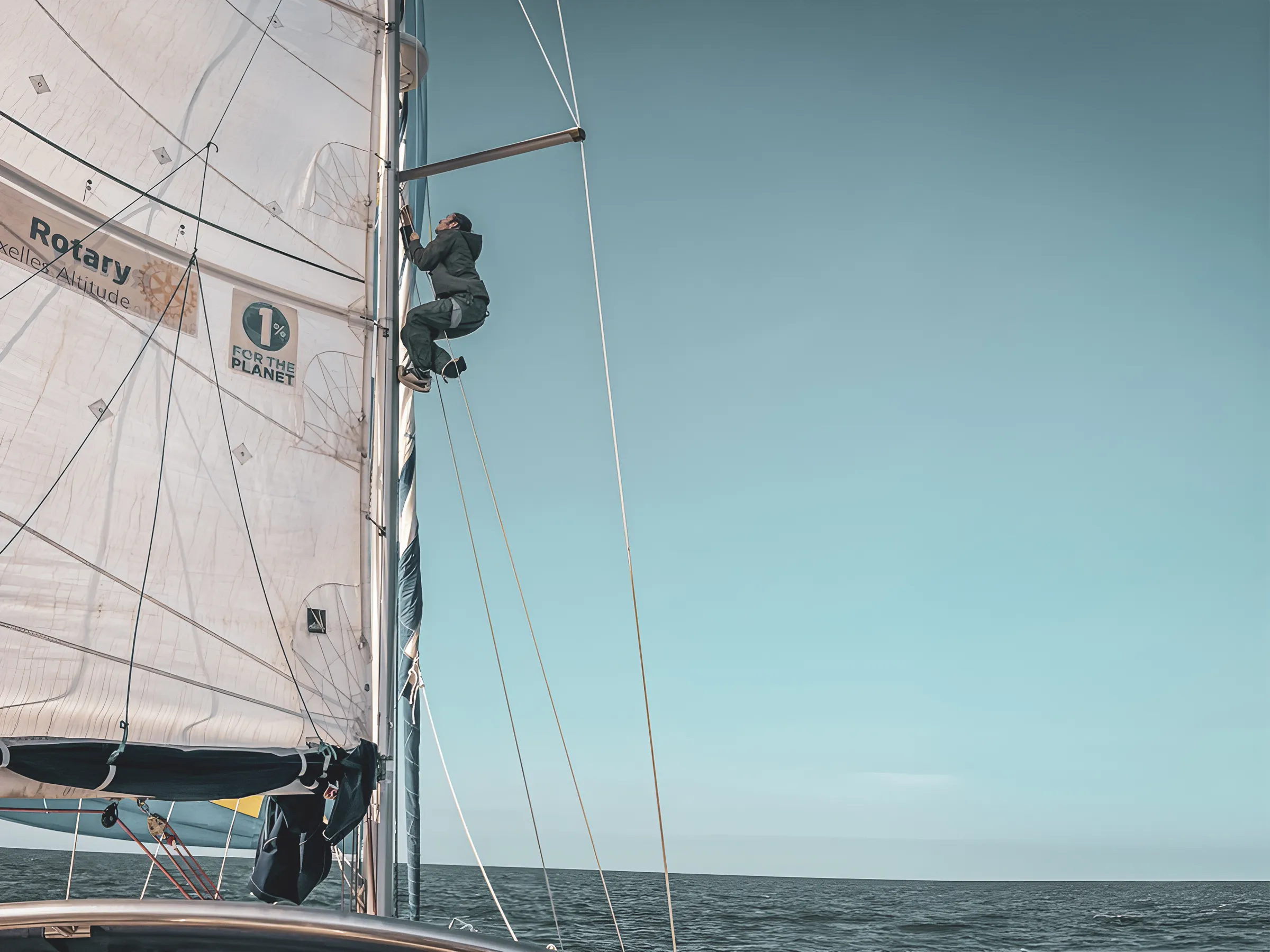 Un marin grimpe sur le mât d’un voilier, sous un ciel bleu, explorant les côtes du Pays de Galle.