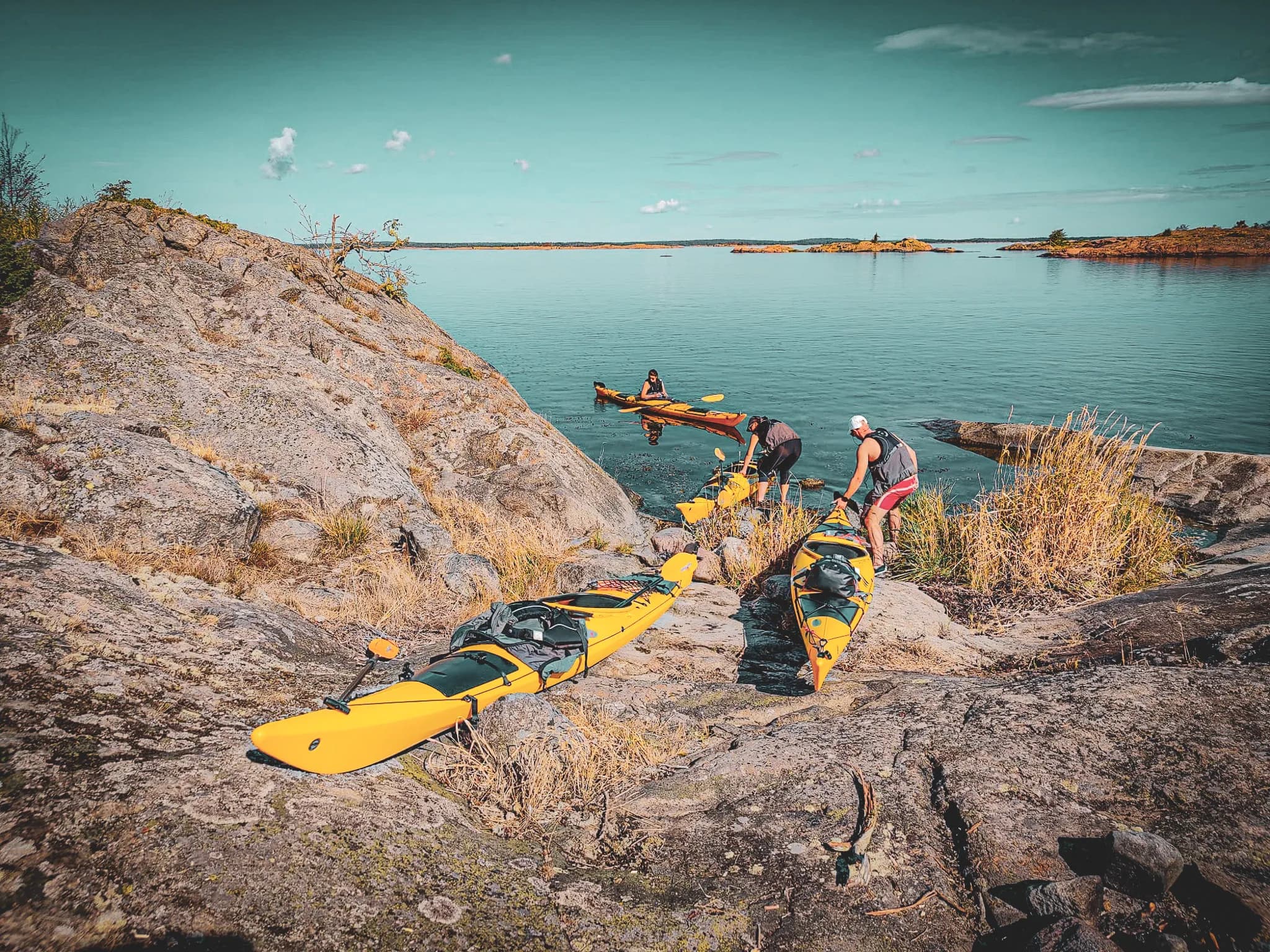 A seaside landscape with yellow kayaks lined up on rocks. Two people are preparing their kayaks while a third is already in the water. The background shows green islets under a