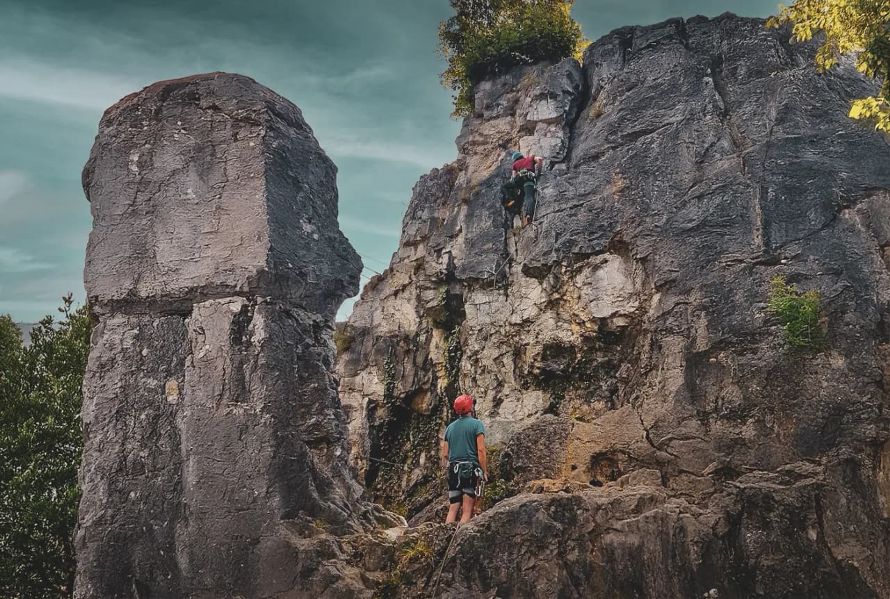 A climbing site with imposing rock formations, where one climber is scaling a vertical wall while another, on the ground, watches.