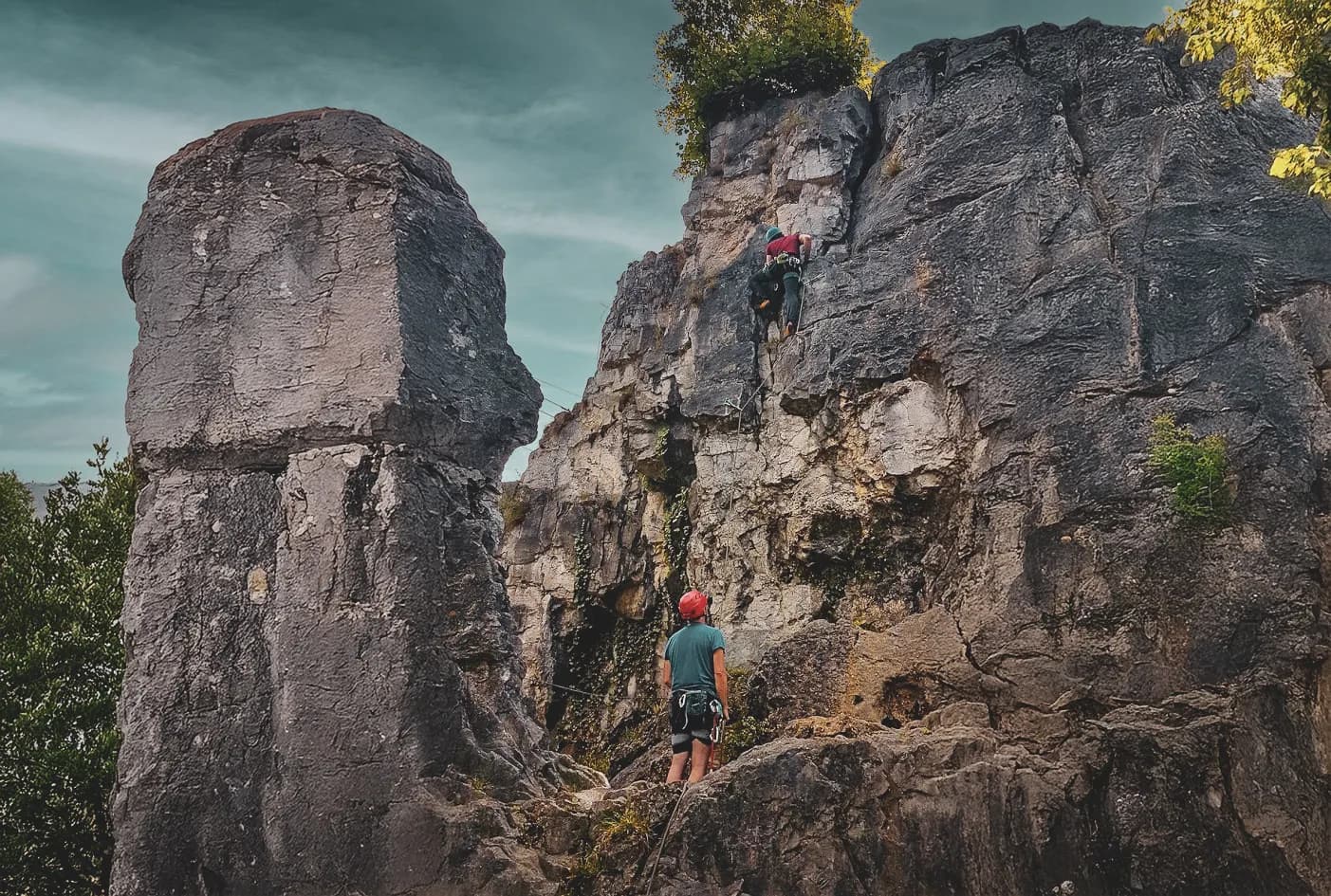 A climbing site with imposing rock formations, where one climber is scaling a vertical wall while another, on the ground, watches. The rocks are covered in moss