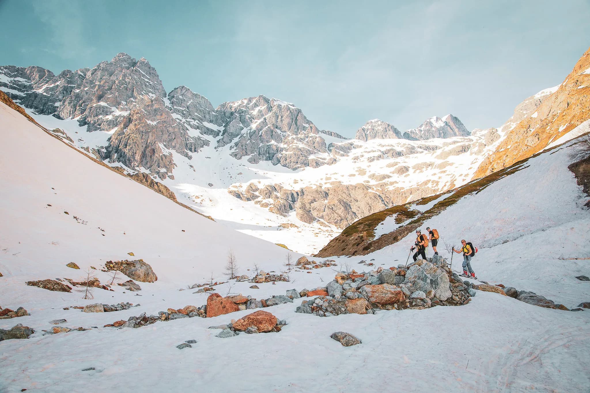 Un paysage montagneux avec des sommets escarpés recouverts de neige, une vallée en pente douce ornée de rochers et de neige. Trois randonneurs équipés de sacs à dos et de bâtons de marche progressent