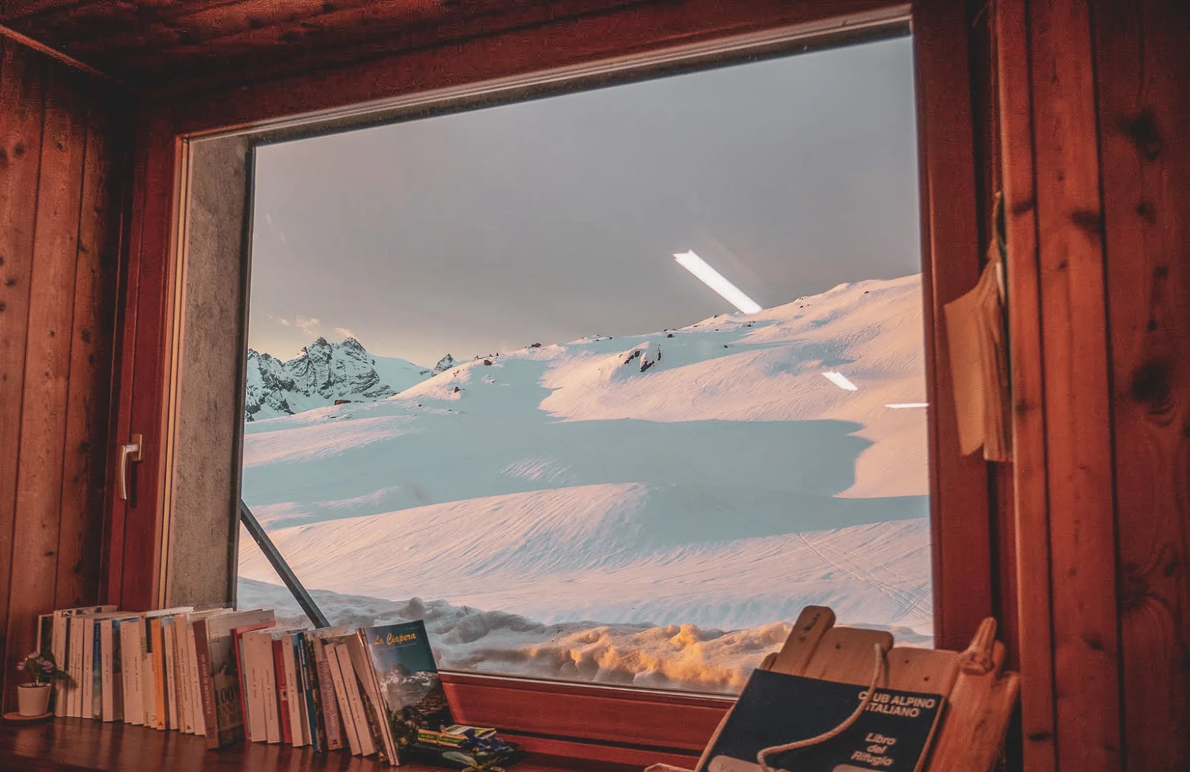 A wooden window offers a panoramic view of a snow-covered mountain landscape. Outside, snow-capped peaks loom under a slightly cloudy sky, with pastel hues hinting at the