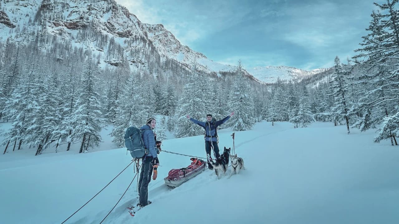 Twee vrolijke wandelaars voor een besneeuwd landschap, met sledehonden en majestueuze bergen.