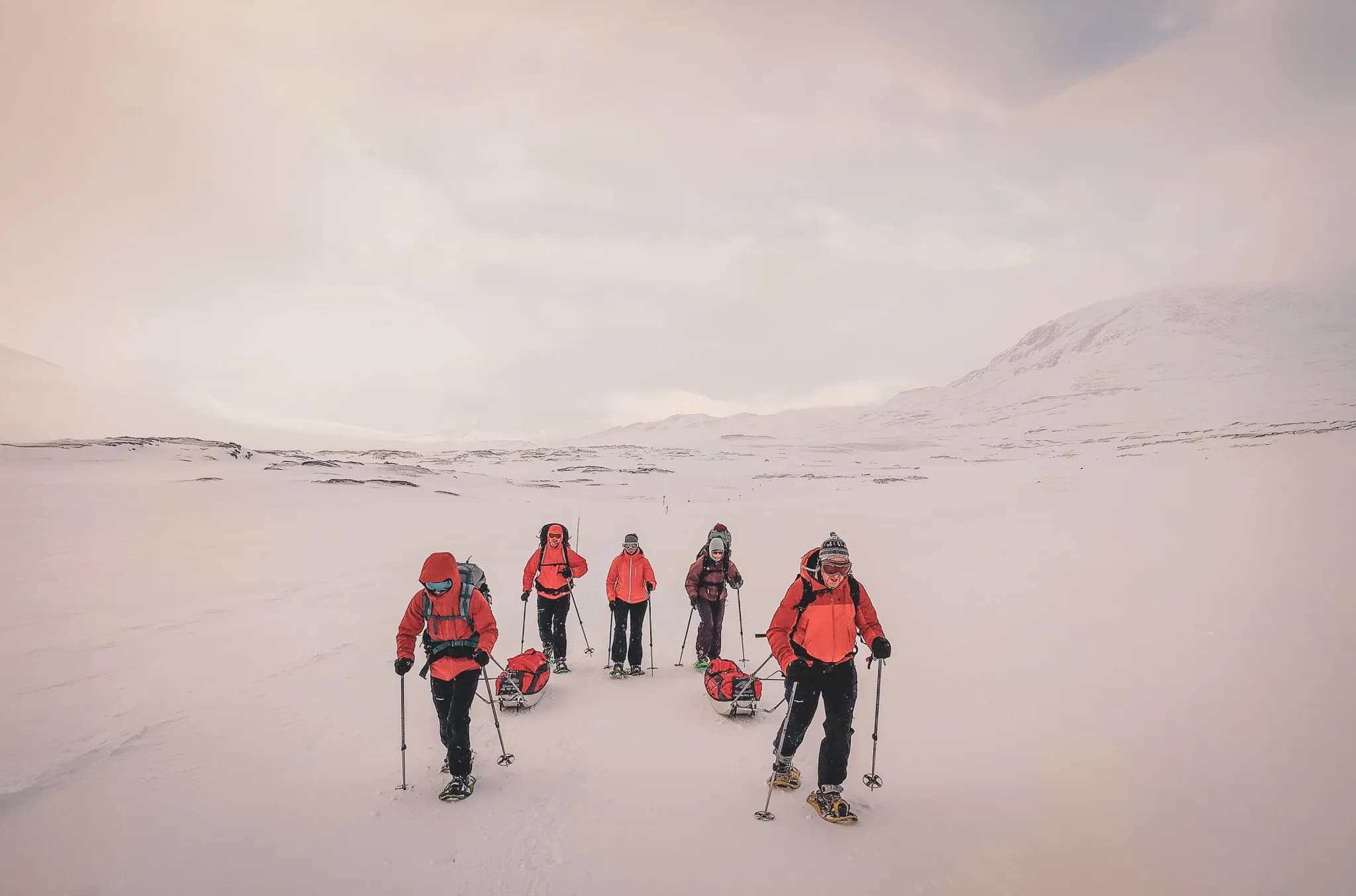 A group of five people on Kungsleden in winter, surrounded by vast expanses of white under cloudy skies. All are wearing warm clothes, mainly bright orange, and ski masks.