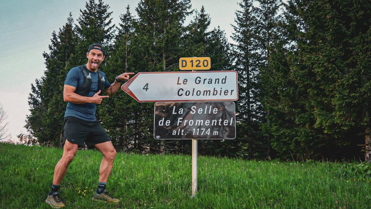 A smiling rider points a signpost to Le Grand Colombier, surrounded by greenery.