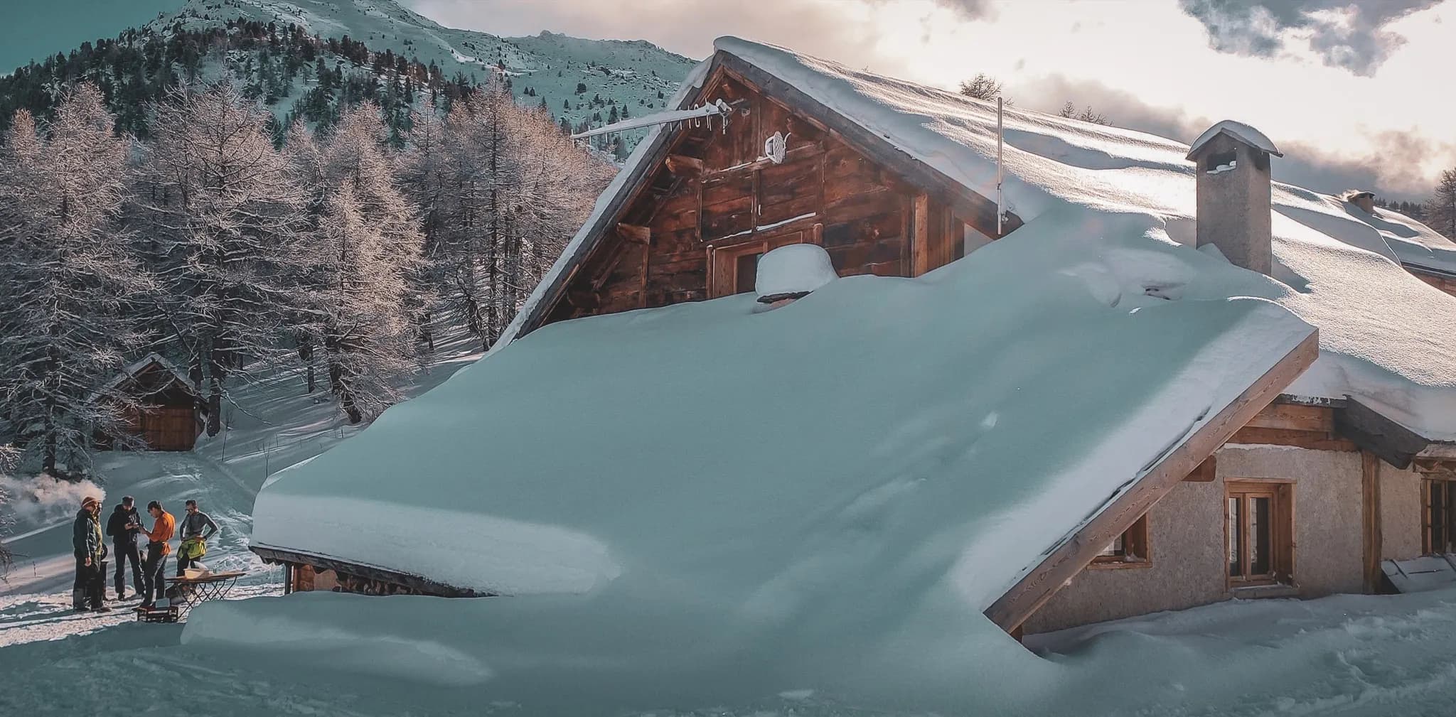 A snow-covered wooden chalet set against a mountain. The scene is peaceful, surrounded by frozen fir trees. A group of people are gathered in front of the chalet, enjoying a convivial moment.