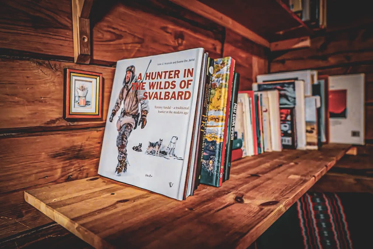 A wooden shelf containing several books, including one in particular entitled "A Hunter in the Wilds of Svalbard". This book is highlighted with an illustration of a hunter in winter clothing, accompanied by a few dogs.
