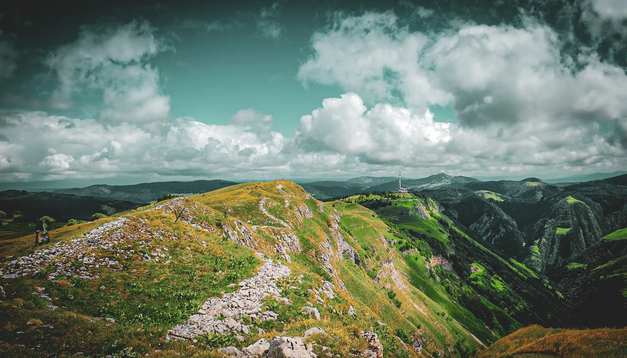 A mountainous landscape with rolling green hills dotted with stones. A winding stone path crosses the scene, while in the distance an aerial stands on a hill. Visit