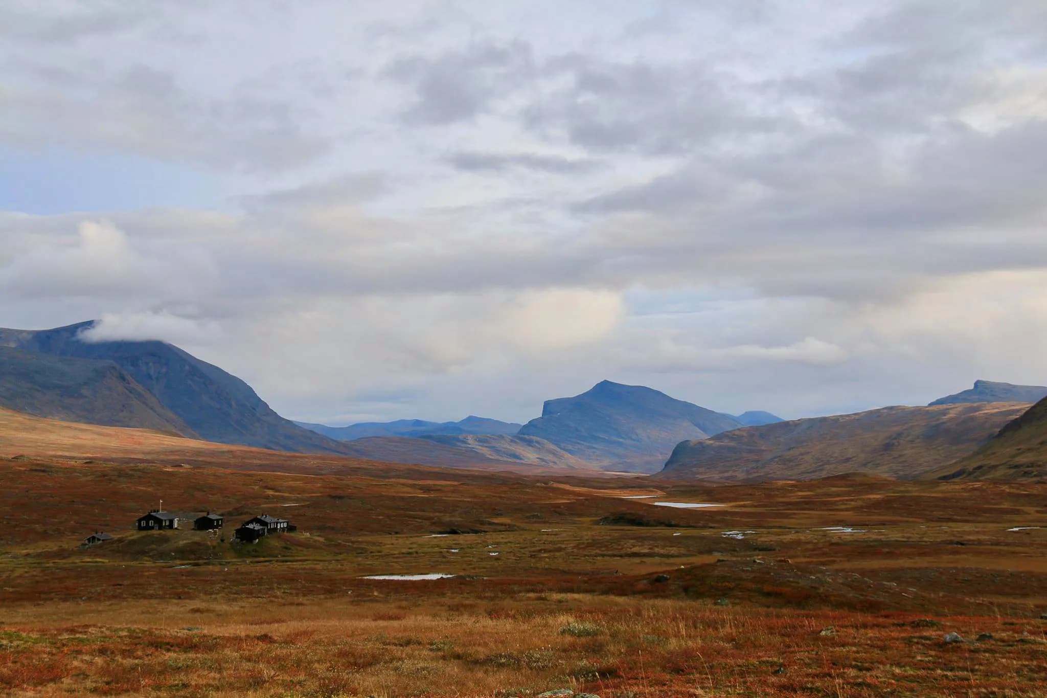 A vast mountain landscape with rolling hills, deep valleys and an overhead cloudy sky. In the foreground, an expanse of land in shades of gold and brown, dotted with a few fla