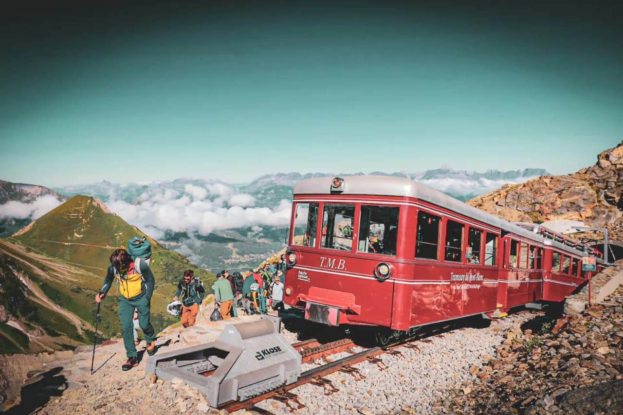 Red train climbing majestic Alpine peaks, surrounded by hikers and clouds.