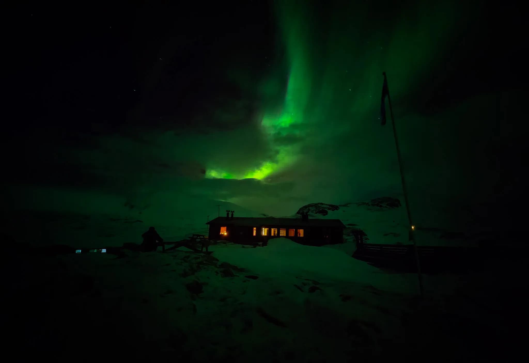 A wooden chalet, an STF refuge, stands in a snowy landscape, illuminated by the light of an aurora borealis that colours the sky in shades of green. Illuminated windows suggest a warm atmosphere inside.