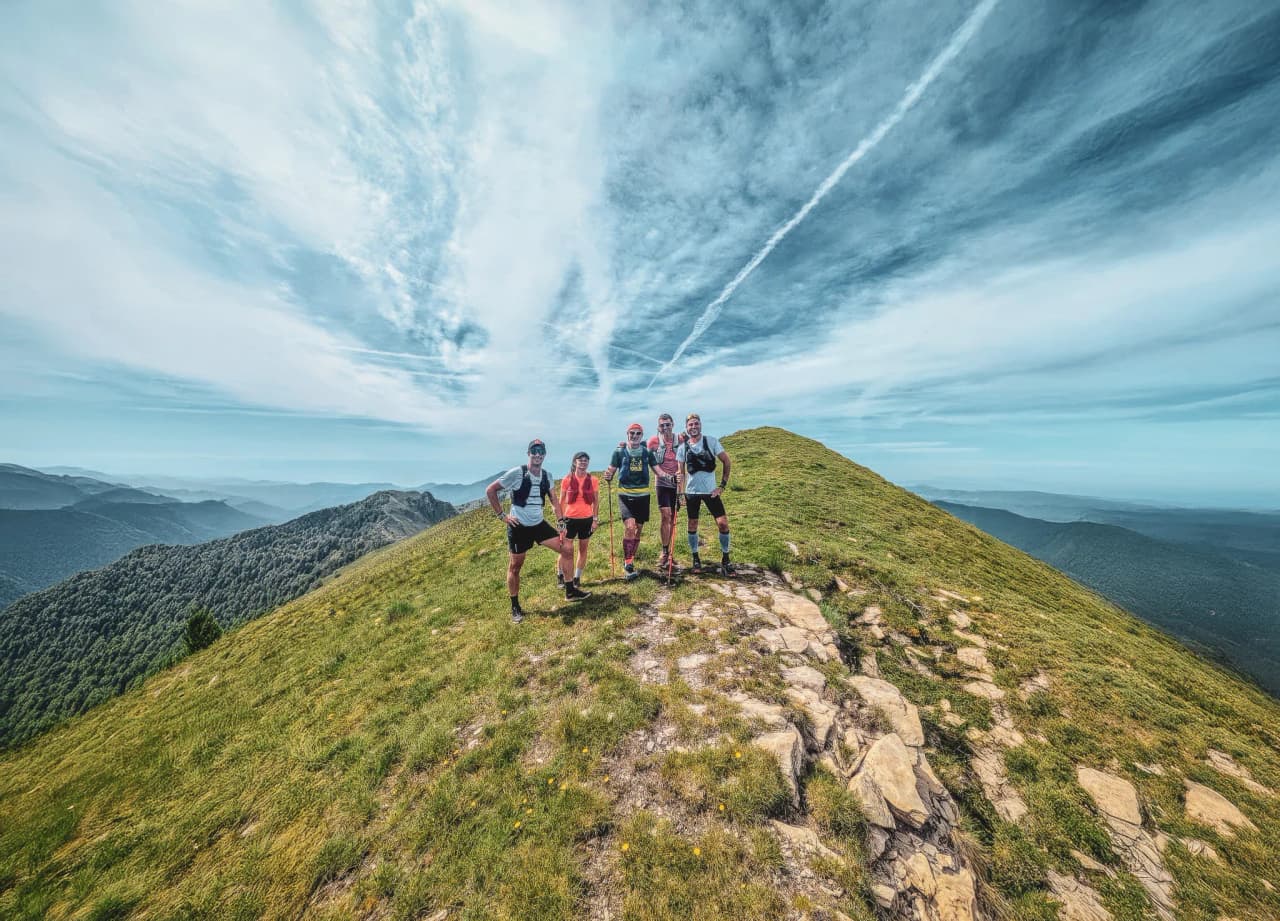 Cinq traileurs souriants au sommet d'une montagne verdoyante, avec un ciel bleu éclatant.