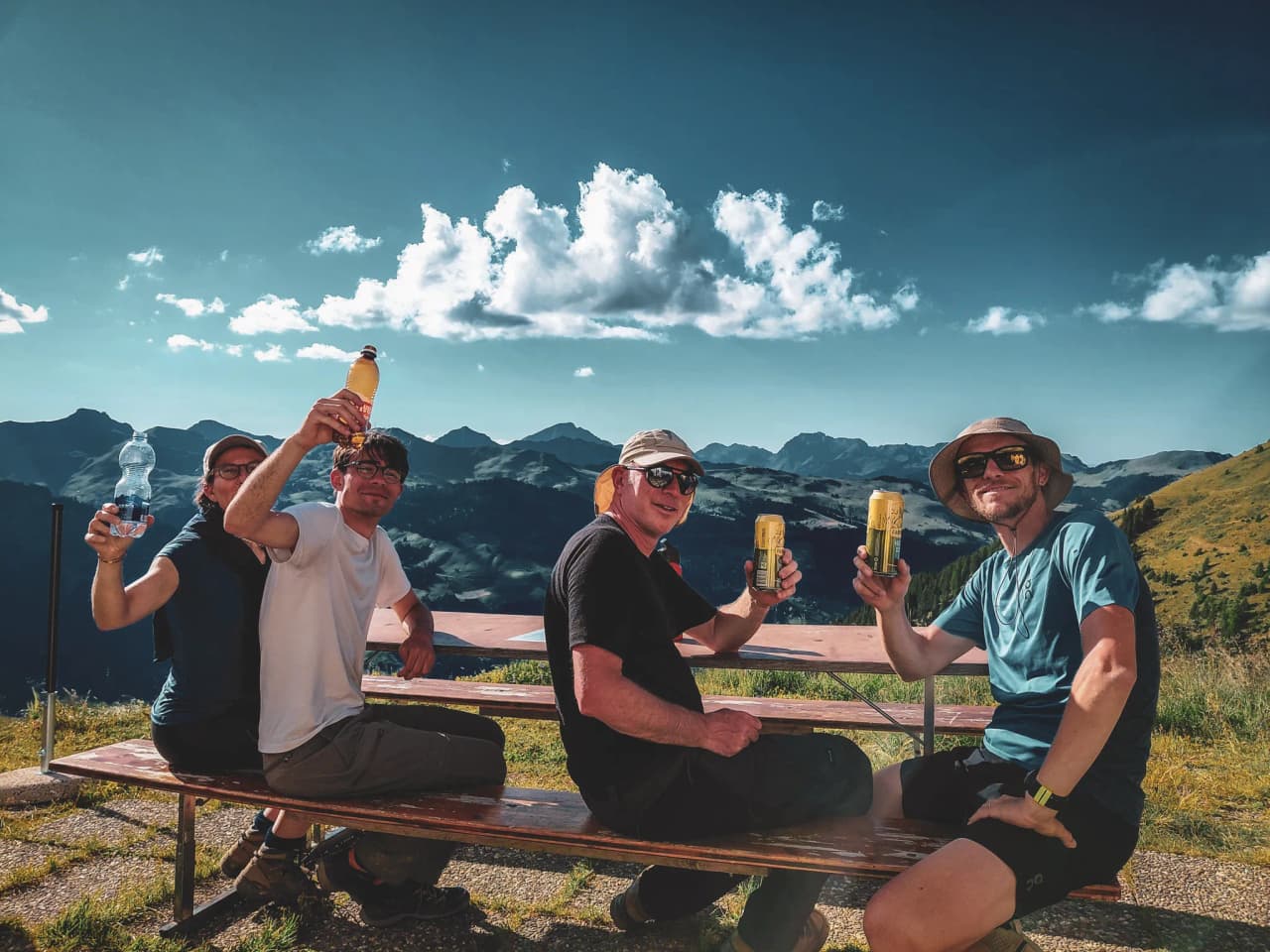Four hikers enjoy a well-earned break with drinks, surrounded by the Alps.