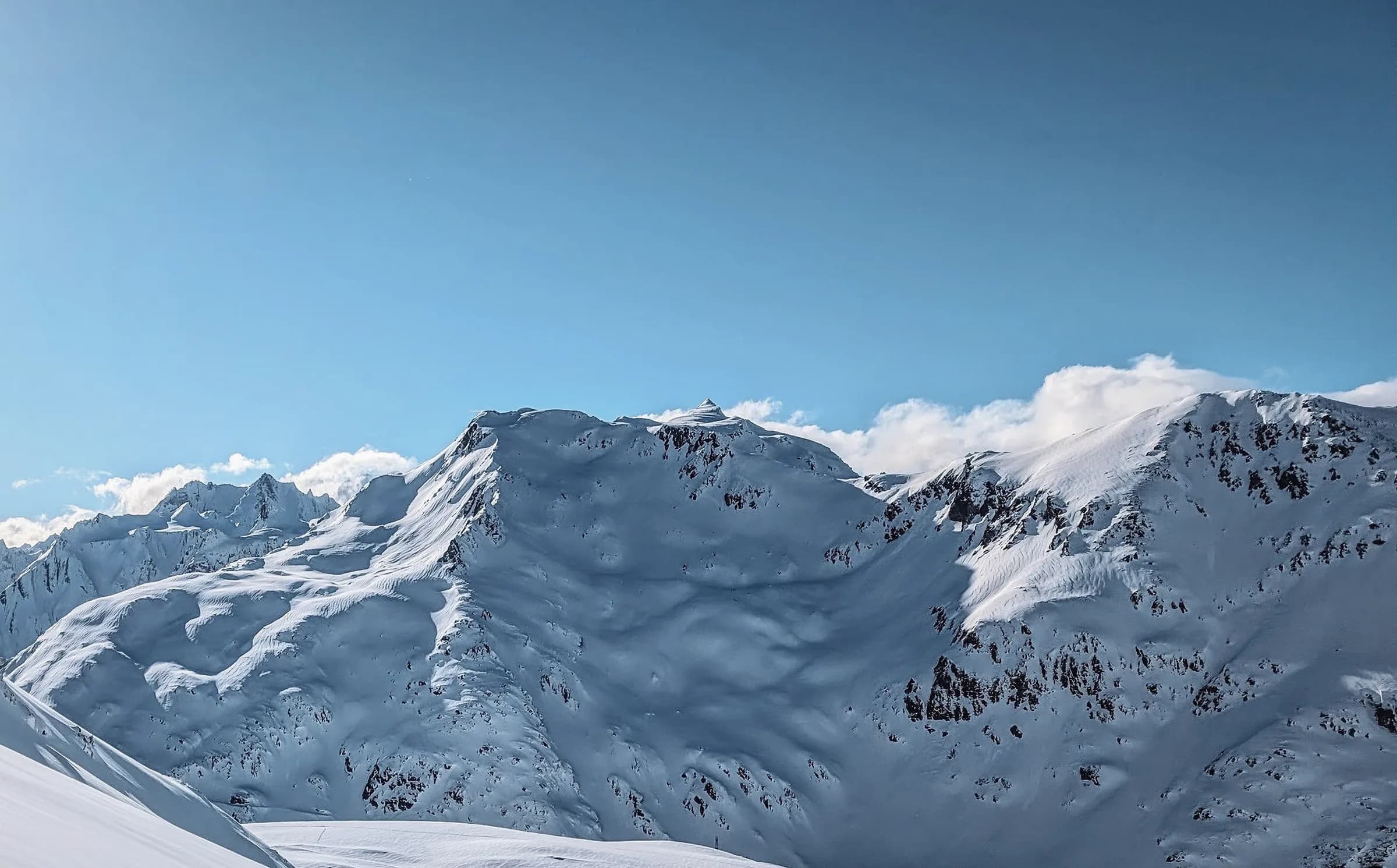 A snow-covered mountain panorama with a clear blue sky and a few clouds. Majestic peaks, covered in a thick layer of snow, rise up to the horizon, creating a serene and peaceful atmosphere.