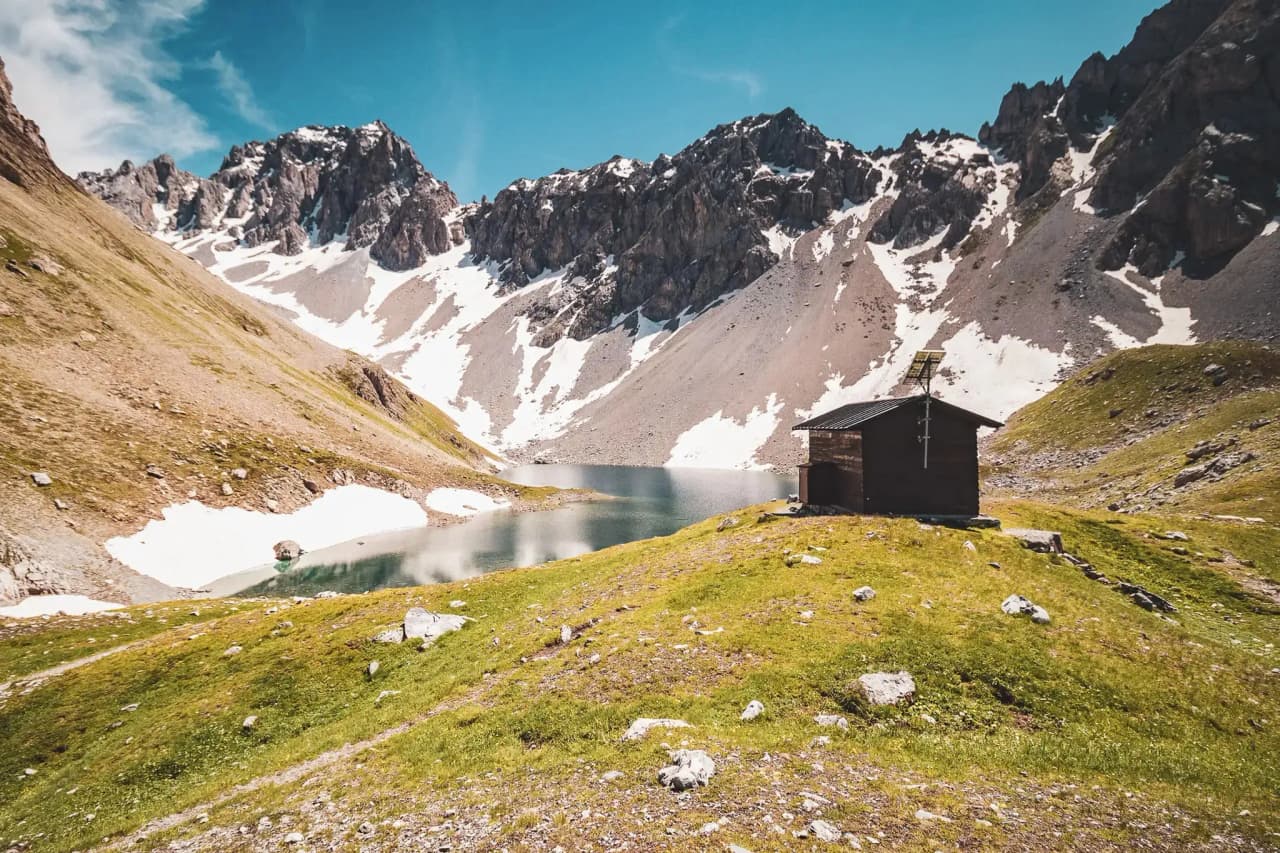 A picturesque chalet on the edge of a lake, surrounded by majestic mountains under a blue sky.