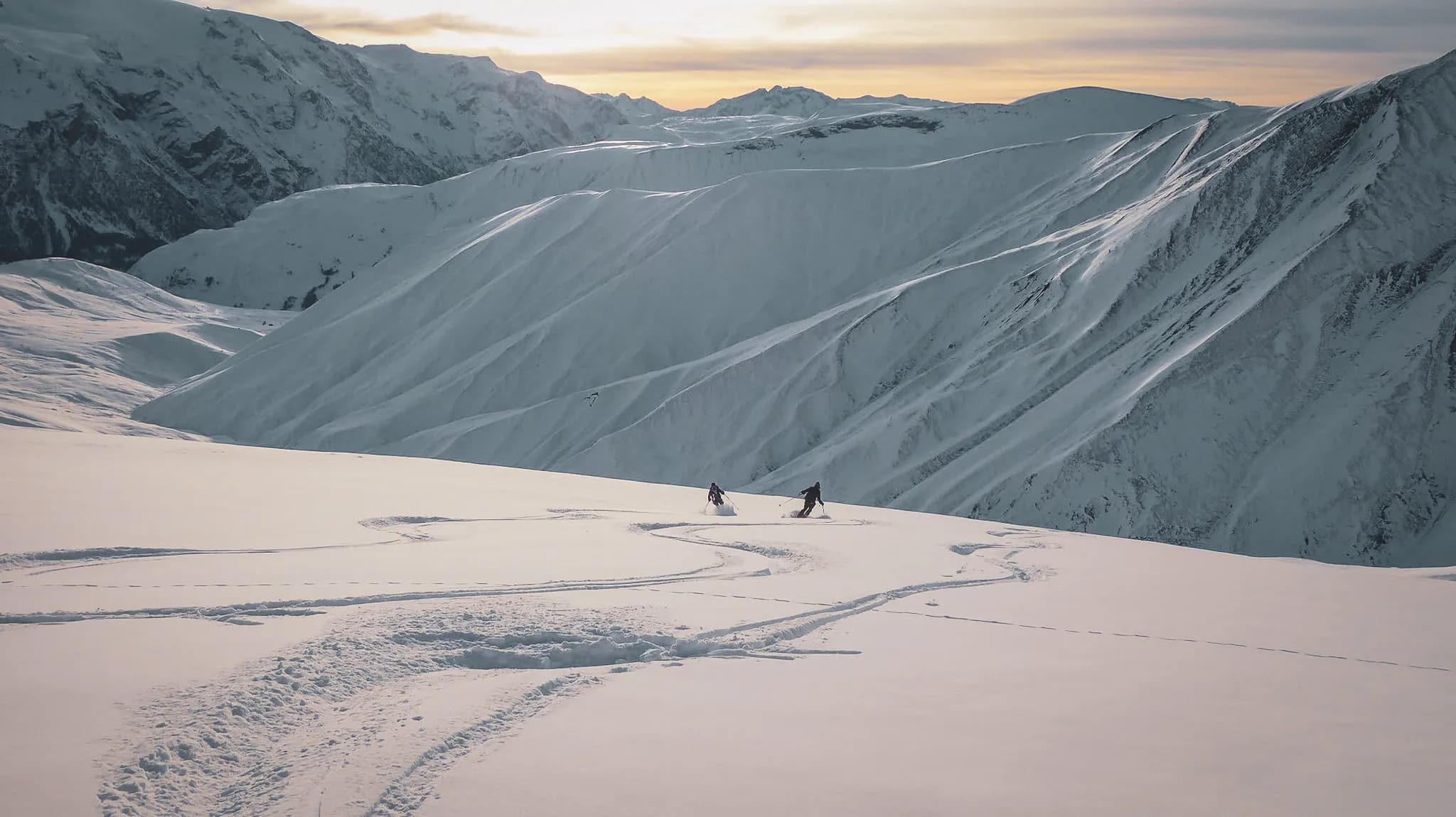 Twee skiërs bewegen door een majestueus alpenlandschap, met besneeuwde toppen op de achtergrond.