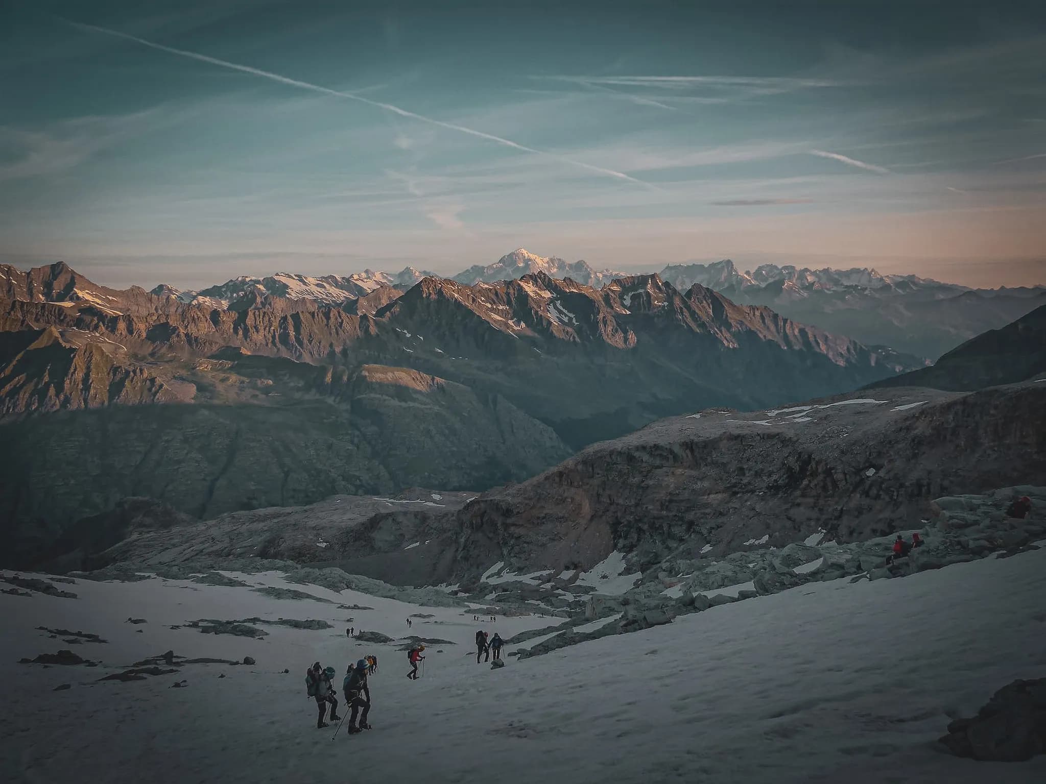 Een berglandschap met majestueuze toppen, waar alpinisten hun weg vinden over besneeuwd en rotsachtig terrein. In de verte bedekken sneeuwvelden de bergpieken, onder een wisselende hemel.