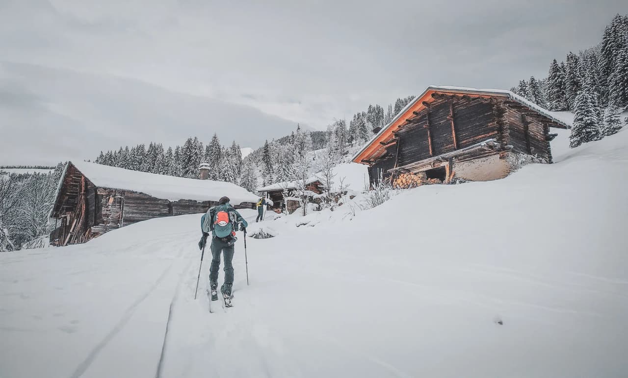A skier walks along a snow-covered path towards wooden chalets set in a wintry landscape. The wooded mountains in the background are dotted with snow-covered trees,