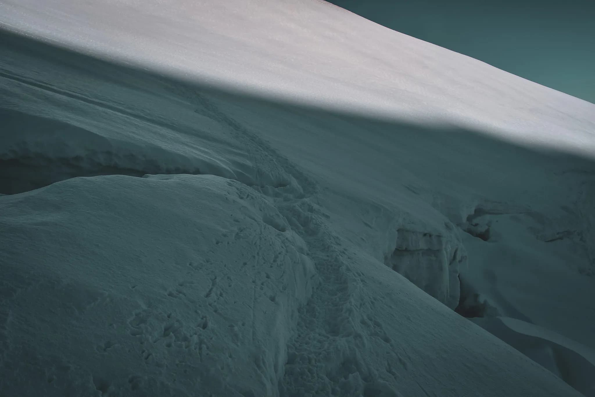 Een besneeuwd landschap met een zachte helling en een ongerept, glad oppervlak. Zachte schaduwen spelen met het licht, terwijl voetsporen door de sneeuw slingeren, teken van recente passage.
