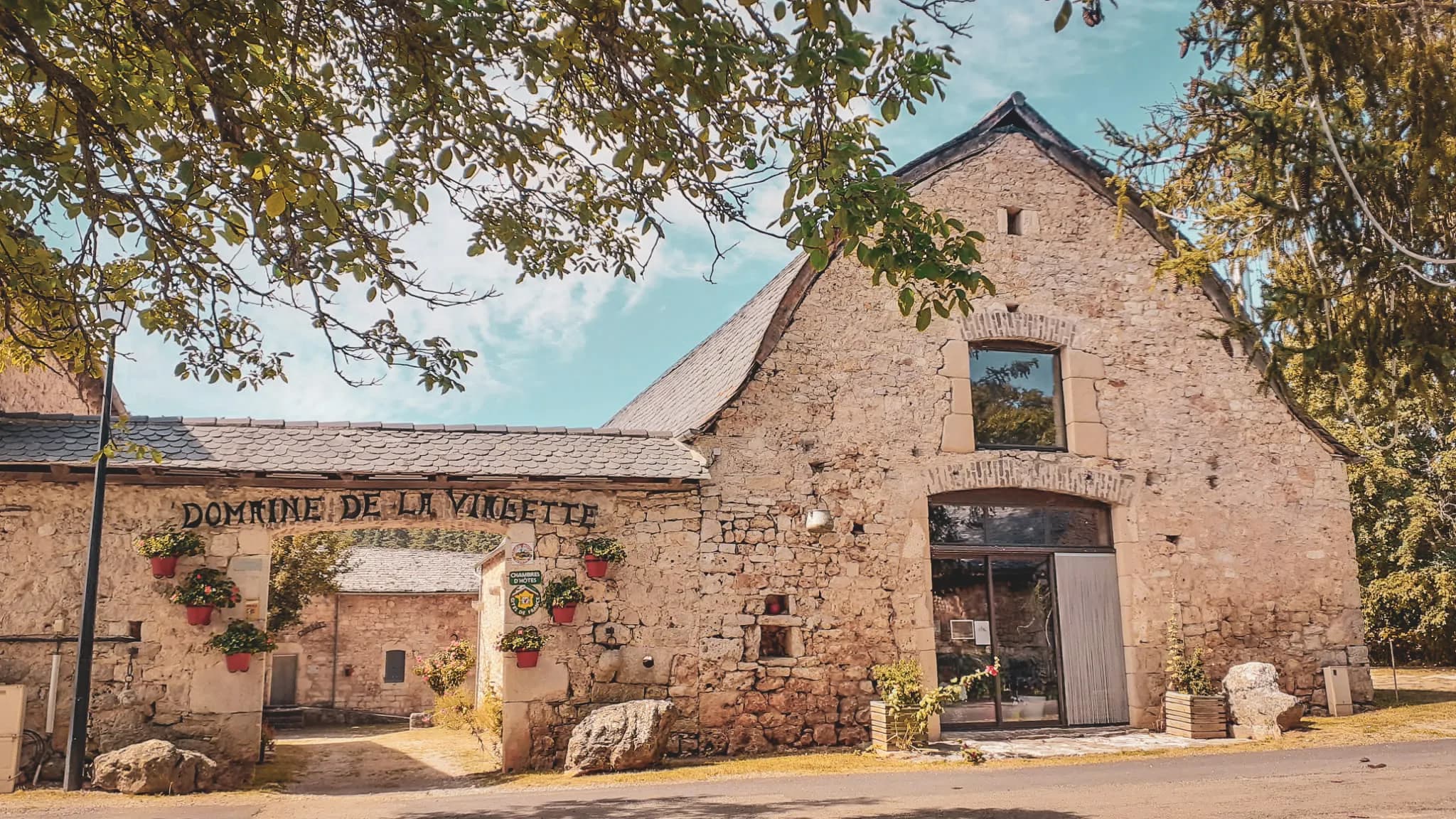 A stone building with a slate roof, decorated with colourful flowers in hanging pots. The inscription "Domaine de la Vignette" can be seen above the entrance. Trees frame the scene