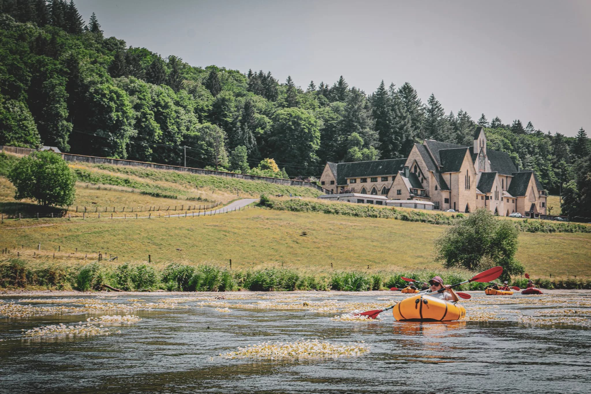 Een kajakker bevaart een rivier, omringd door weelderig groen met bloemen die op het wateroppervlak drijven. Op de achtergrond staat een groot gebouw met een opvallende bouwstijl, waarschijnlijk een hotel of hostel.