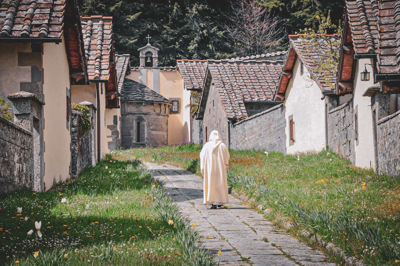 A monk walking through the Camaldoli Monastery, with stone walls on either side and a lawn filled with colourful flowers.
