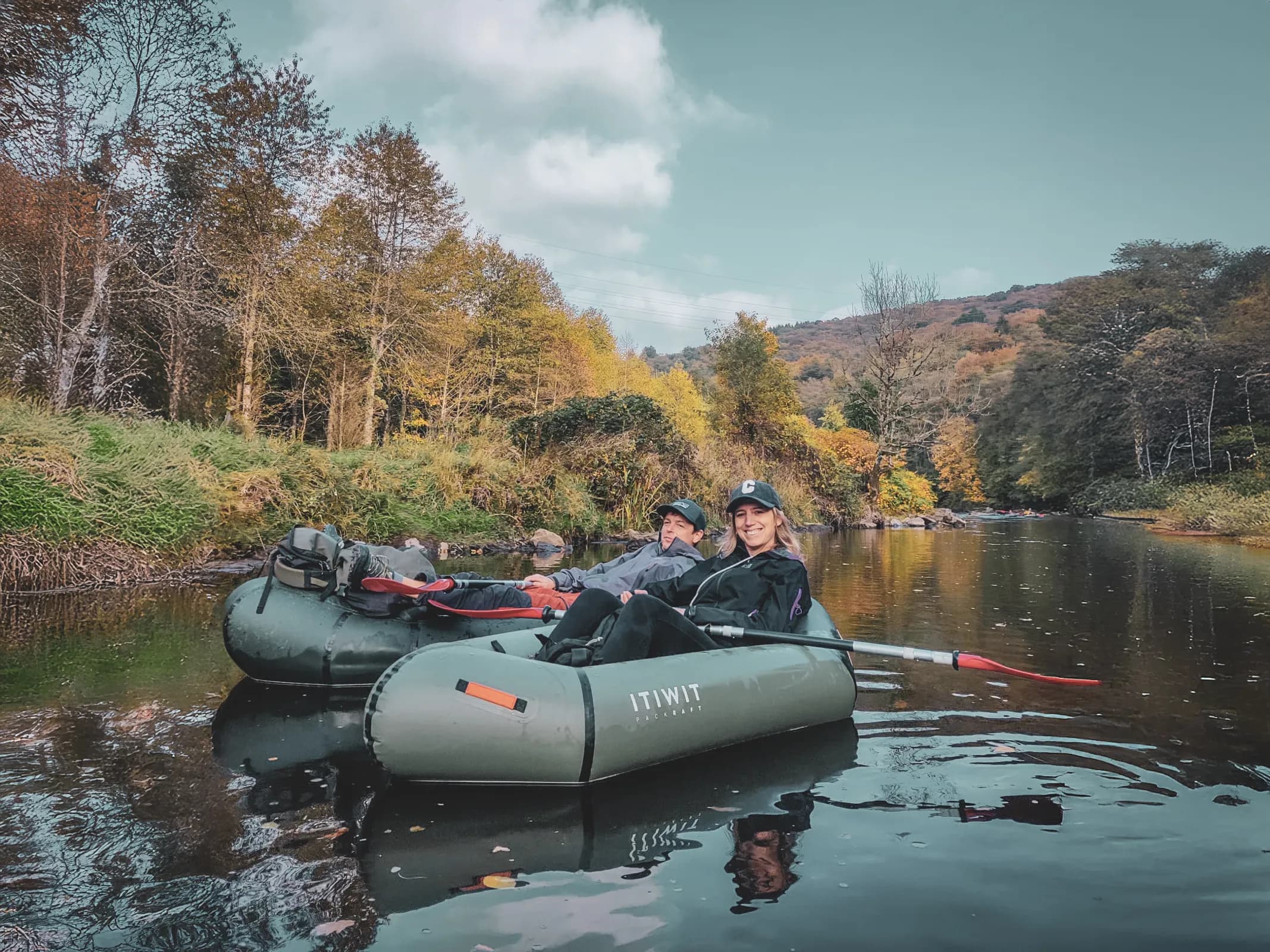 Two people enjoying a packraft ride on a river lined with colourful trees in autumn.