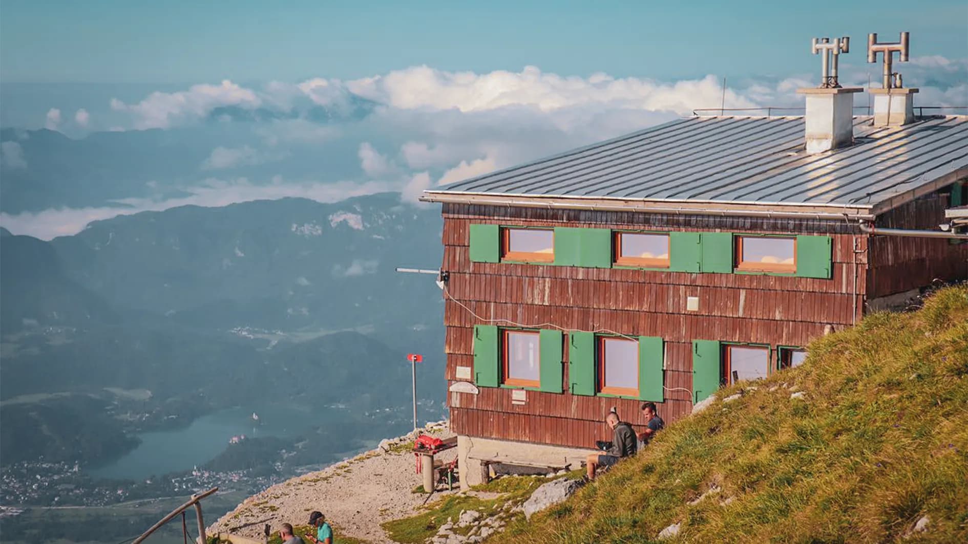 A wooden chalet with green shutters, set high above a mountain valley. The surrounding landscape is made up of green hills and clouds rising above the mountains. Close to the