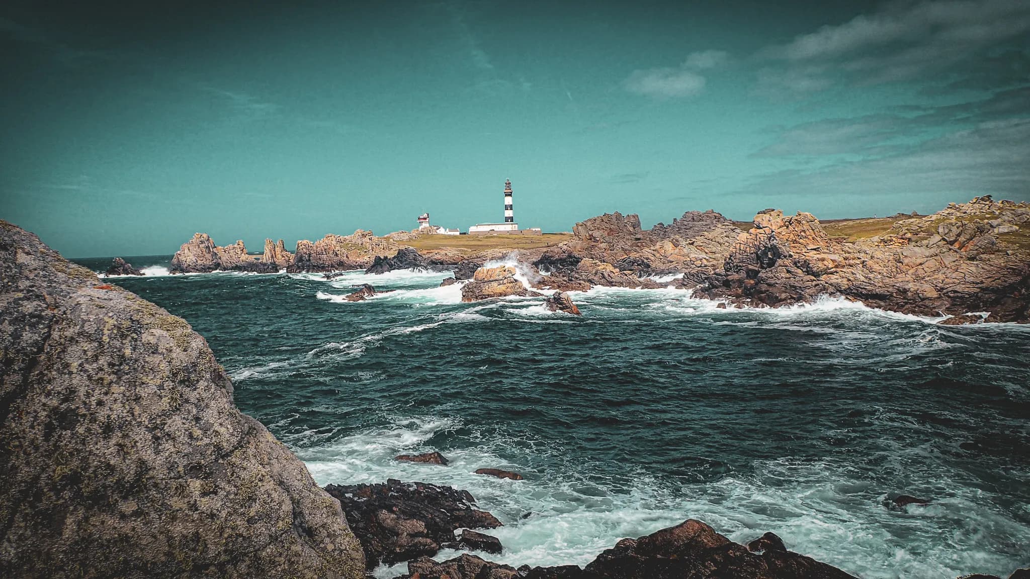 A black and white lighthouse stands on a small rocky island, surrounded by tumultuous ocean waves. Large rock formations emerge from the water, while splashes of water form on the horizon.